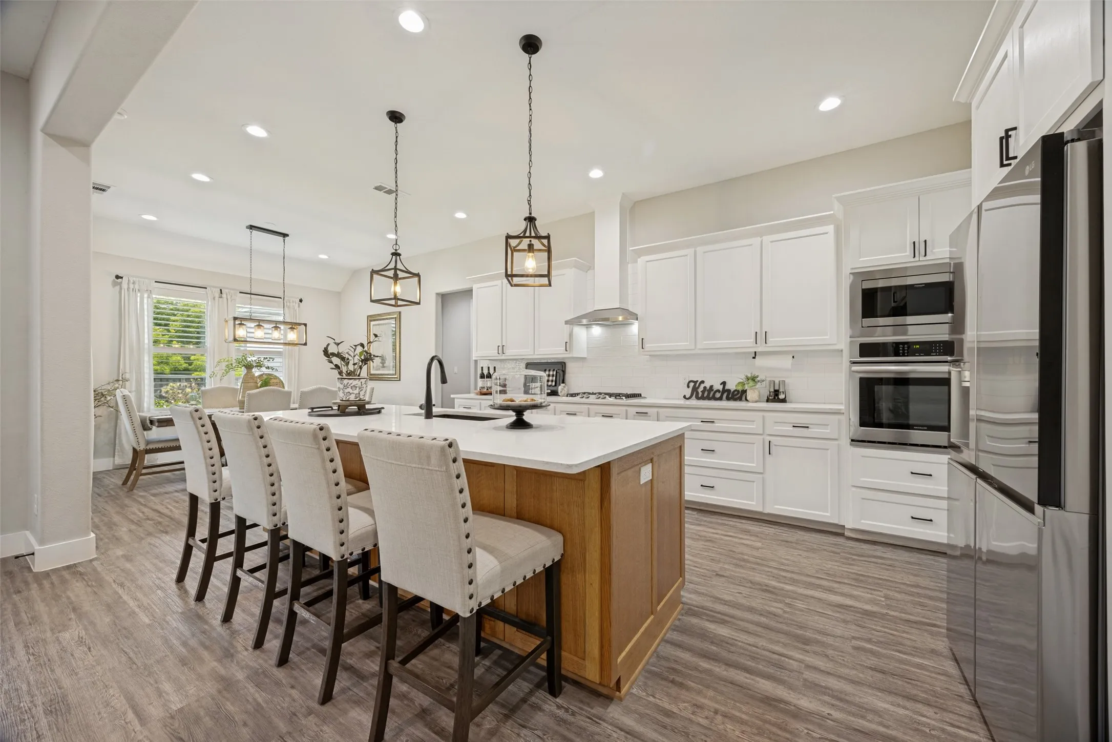 Open-concept kitchen featuring an oversized island with seating, wood-finish flooring, white cabinetry, integrated stainless steel appliances, and ceiling-mounted light fixtures