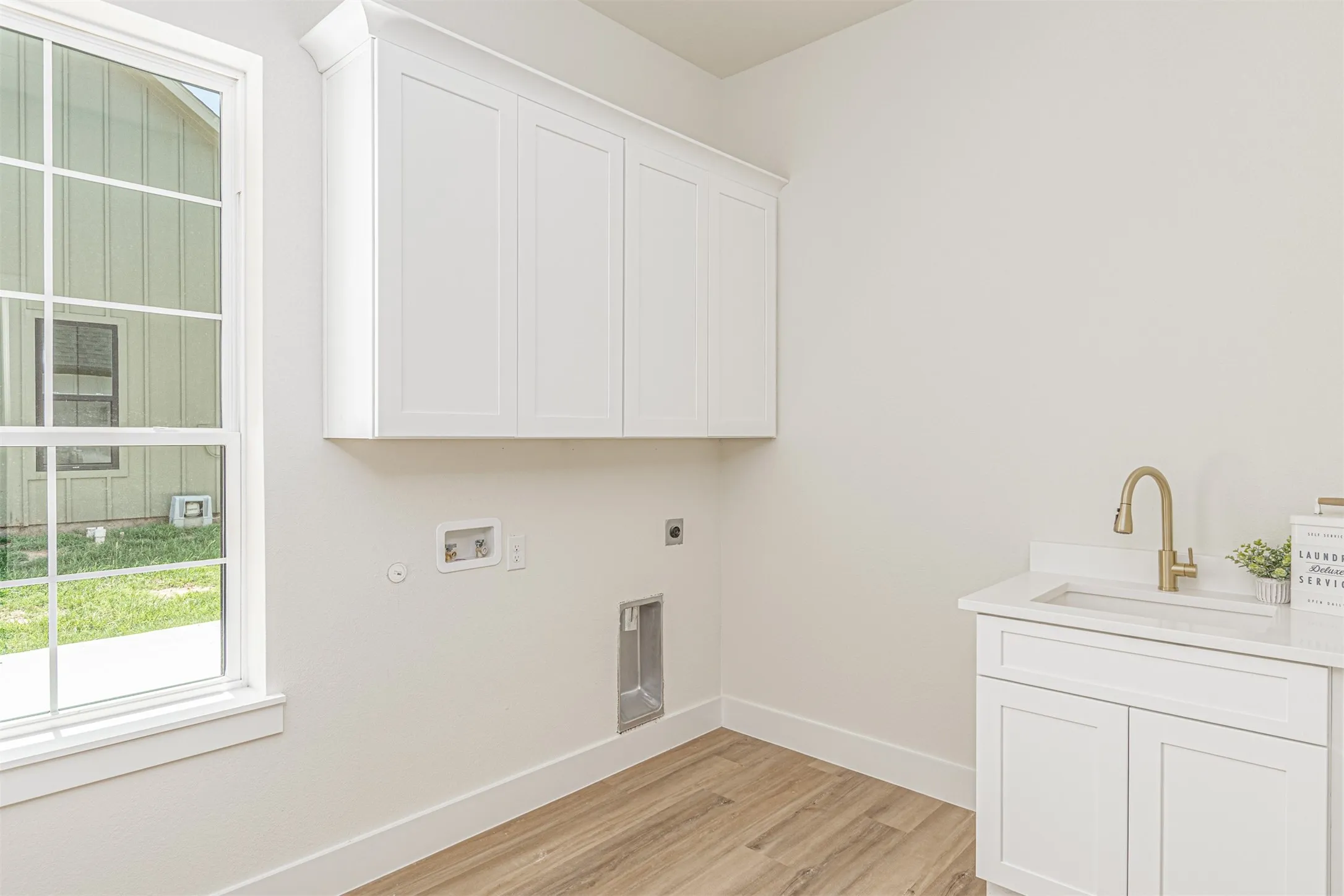 Laundry room featuring white cabinetry, a built-in sink with a gooseneck faucet, wood-finish flooring, and a large window