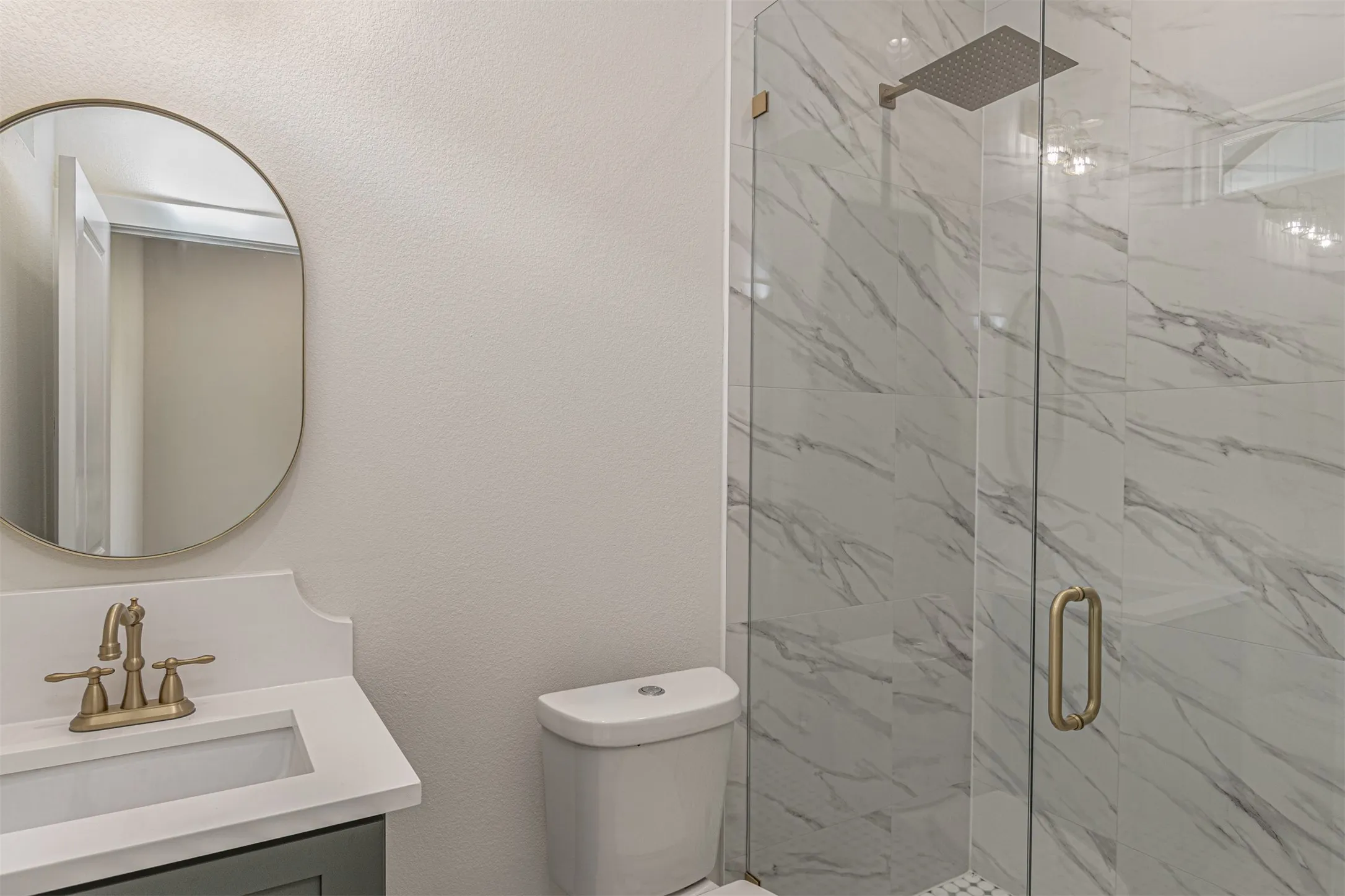 Bathroom featuring a walk-in shower with clear glass enclosure and marble-look tile walls, a rain showerhead, a vanity with a white countertop and undermount sink, a bronze-finish faucet, and an oval mirror