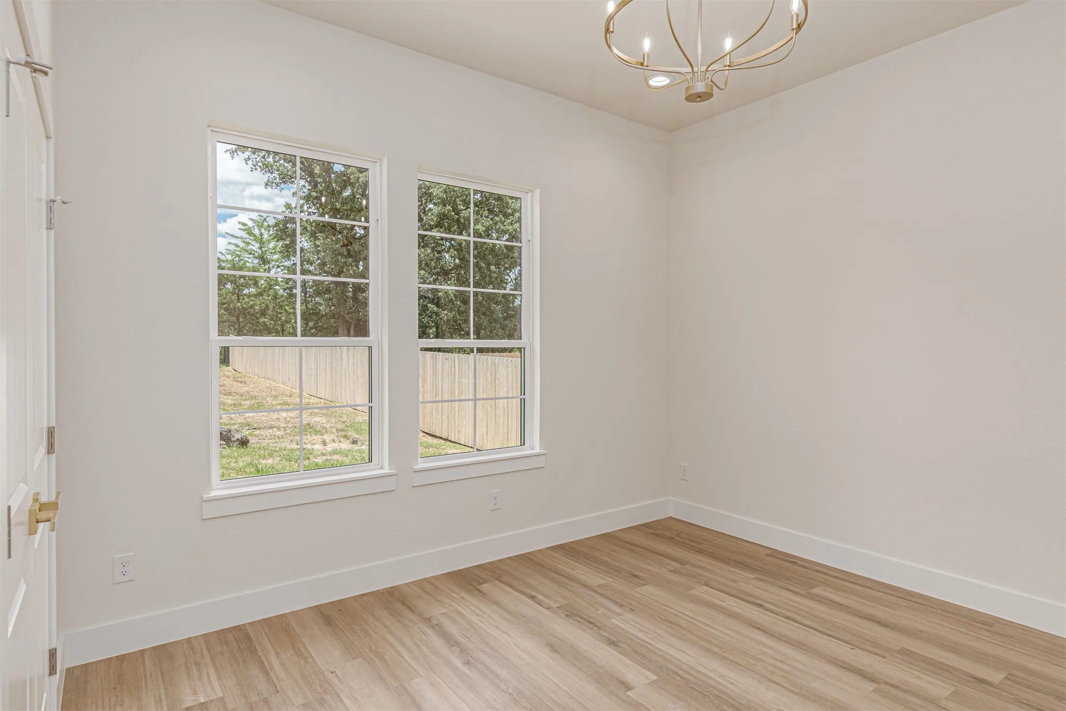 Room featuring two double-hung windows, wood-finish flooring, a contemporary chandelier, white baseboards, and a paneled interior door with brass hardware