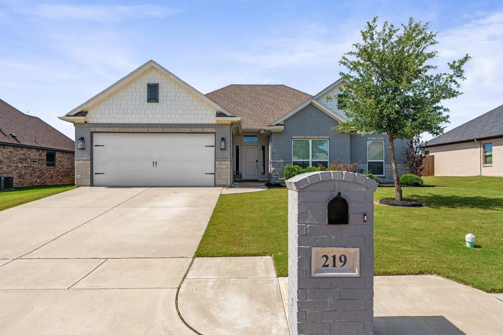 View of front of home, gray brick painted mailbox, 2 car garage and driveway. Gray painted brick exterior around house with white painted shingle style siding.