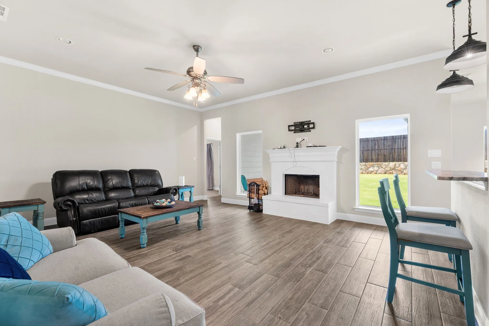 Living Room with ceiling fan, white painted brick wood burning fire place, windows for natural lighting, brown/gray wood tile flooring. Crown Moulding.