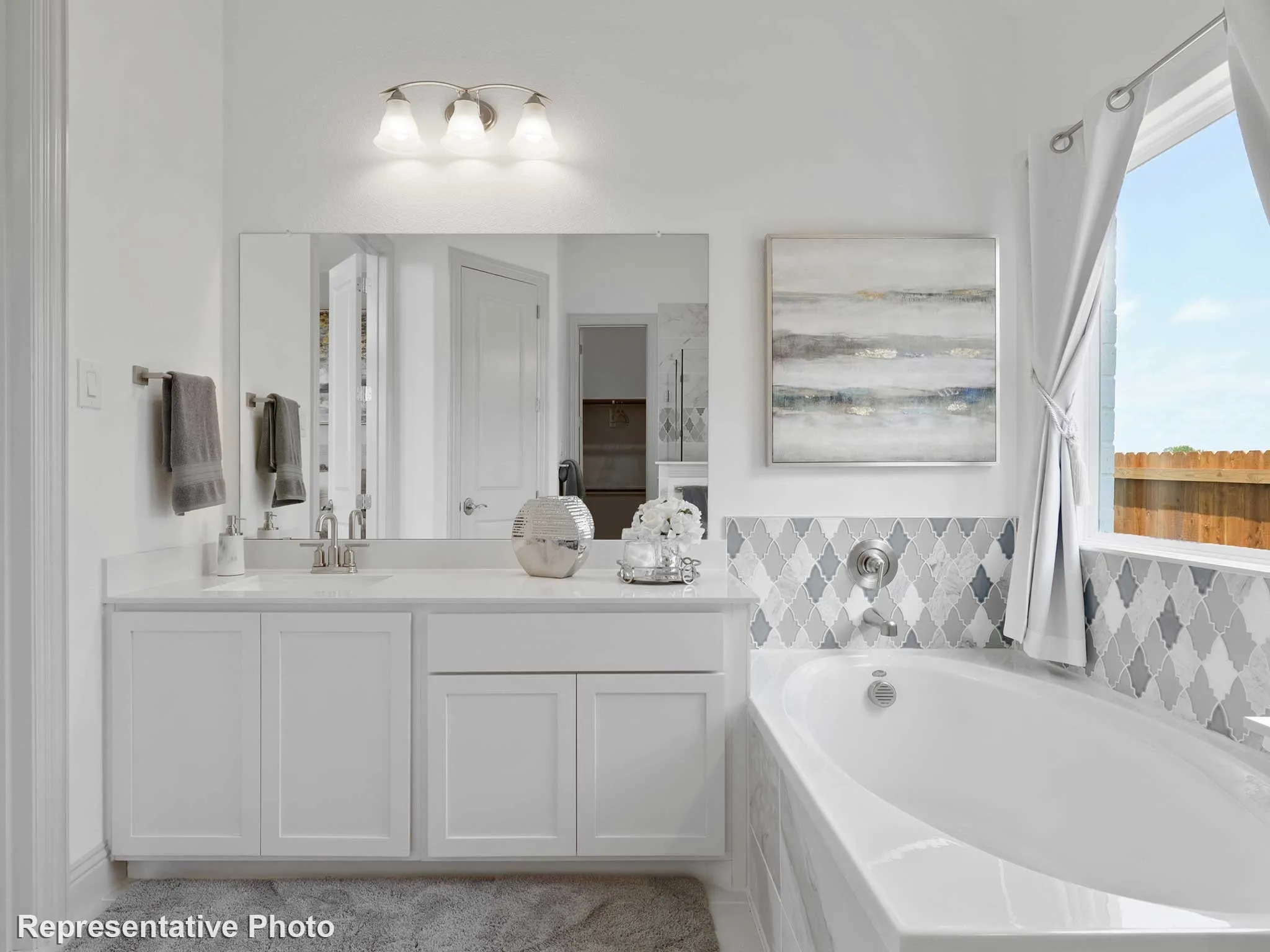 White bathroom featuring a built-in tub with decorative tile surround, a single vanity with a quartz-style countertop, and a large mirror with overhead light fixture
