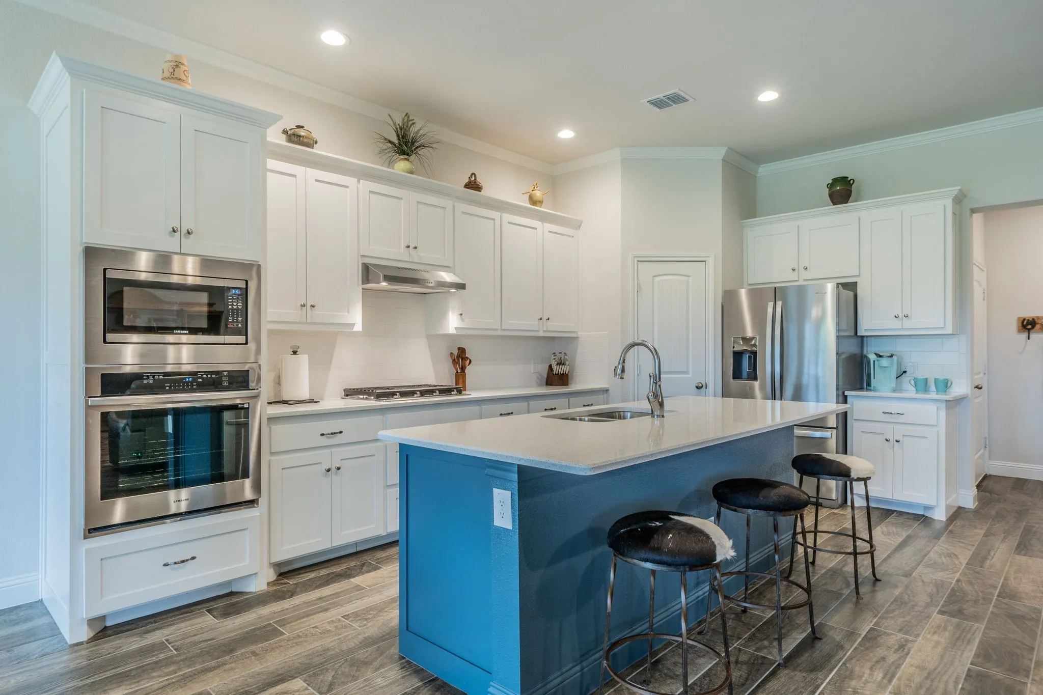 Contemporary kitchen featuring a blue island with seating, stainless steel appliances, white cabinetry, recessed lighting, and wood-finish tile flooring