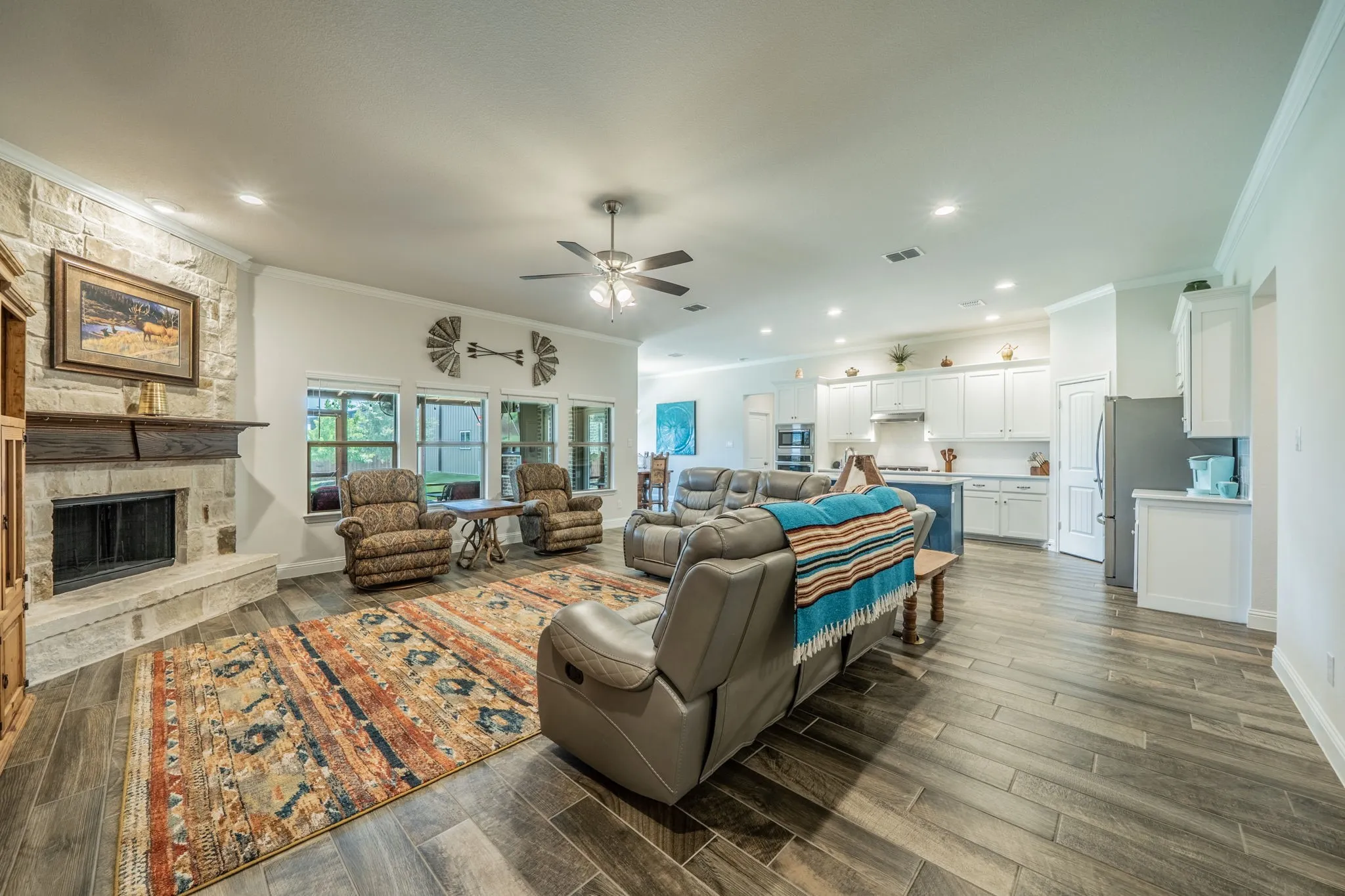 Open-concept living space featuring wood-finish tile flooring, a stone fireplace with a substantial wood mantel, and multiple windows providing natural light