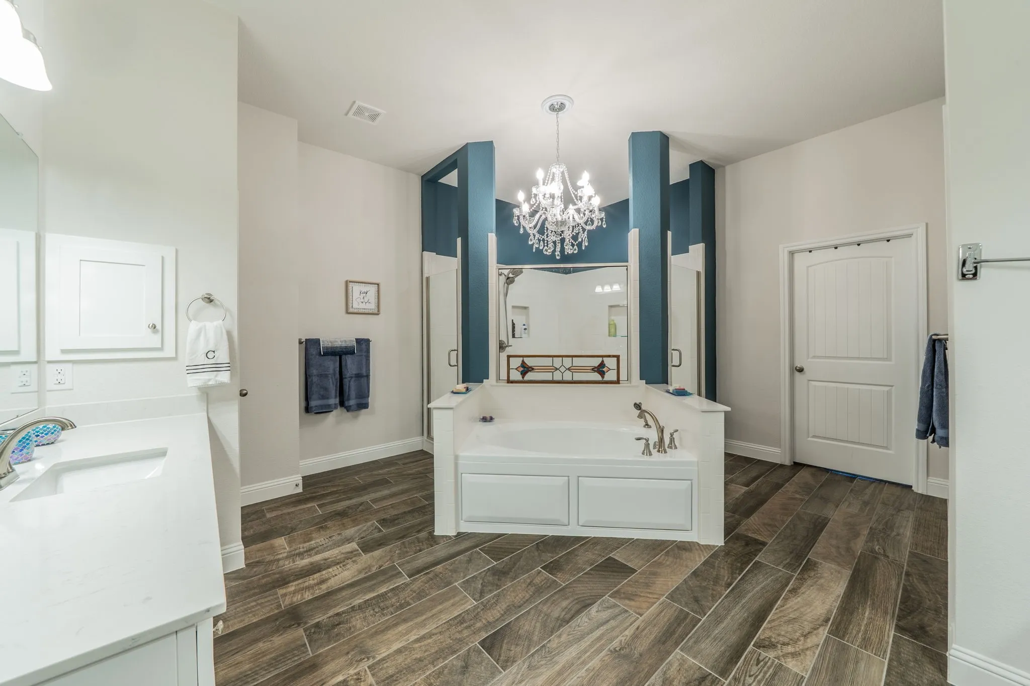 Spacious Master bathroom featuring a built-in soaking tub, wood-finish tile flooring, dual vanities with white countertops, and a decorative chandelier