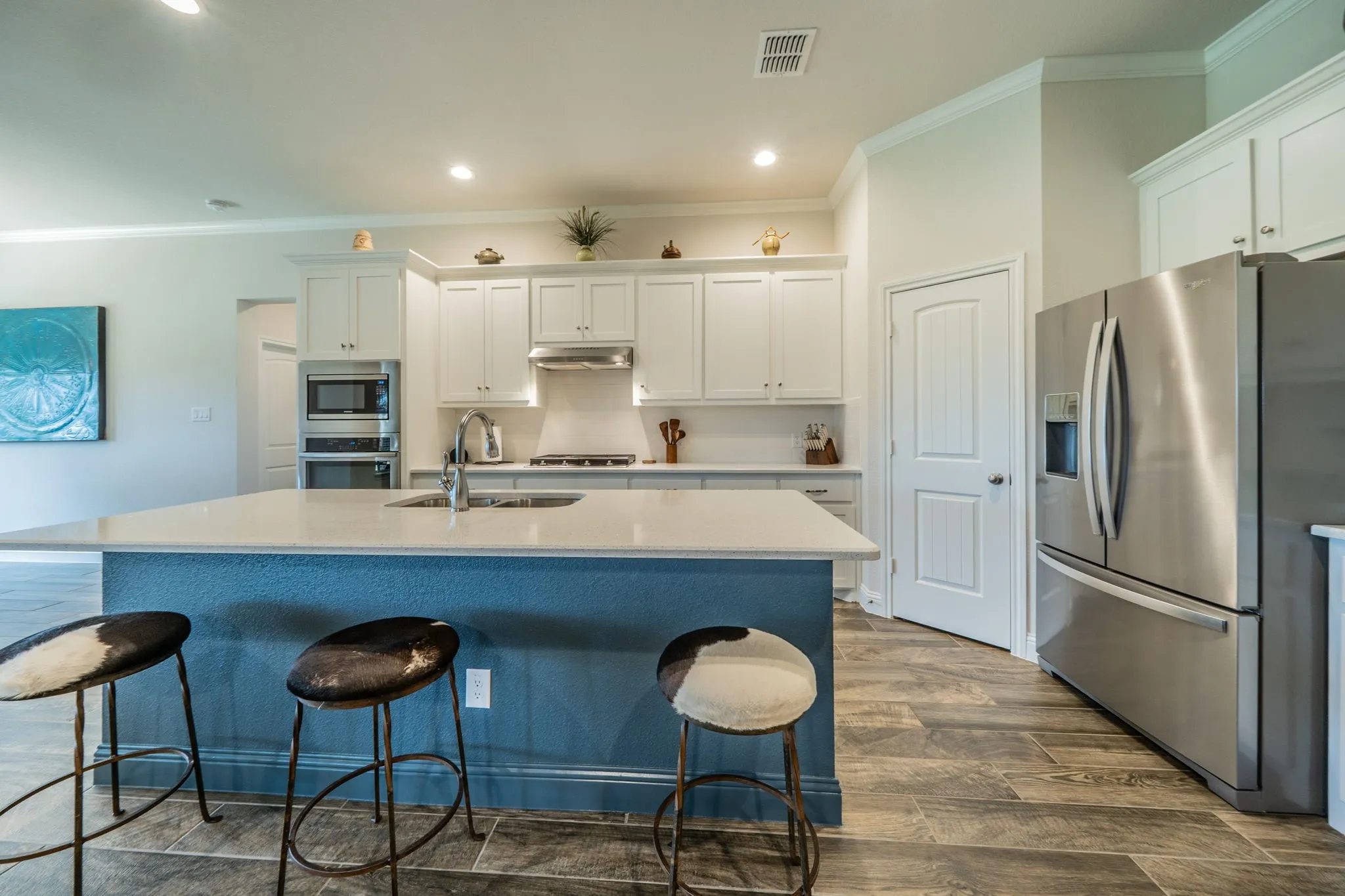 Kitchen featuring a central island with a light-colored countertop and a blue-painted base, white cabinetry, integrated stainless steel appliances, a tile backsplash, and wood-finish tile flooring