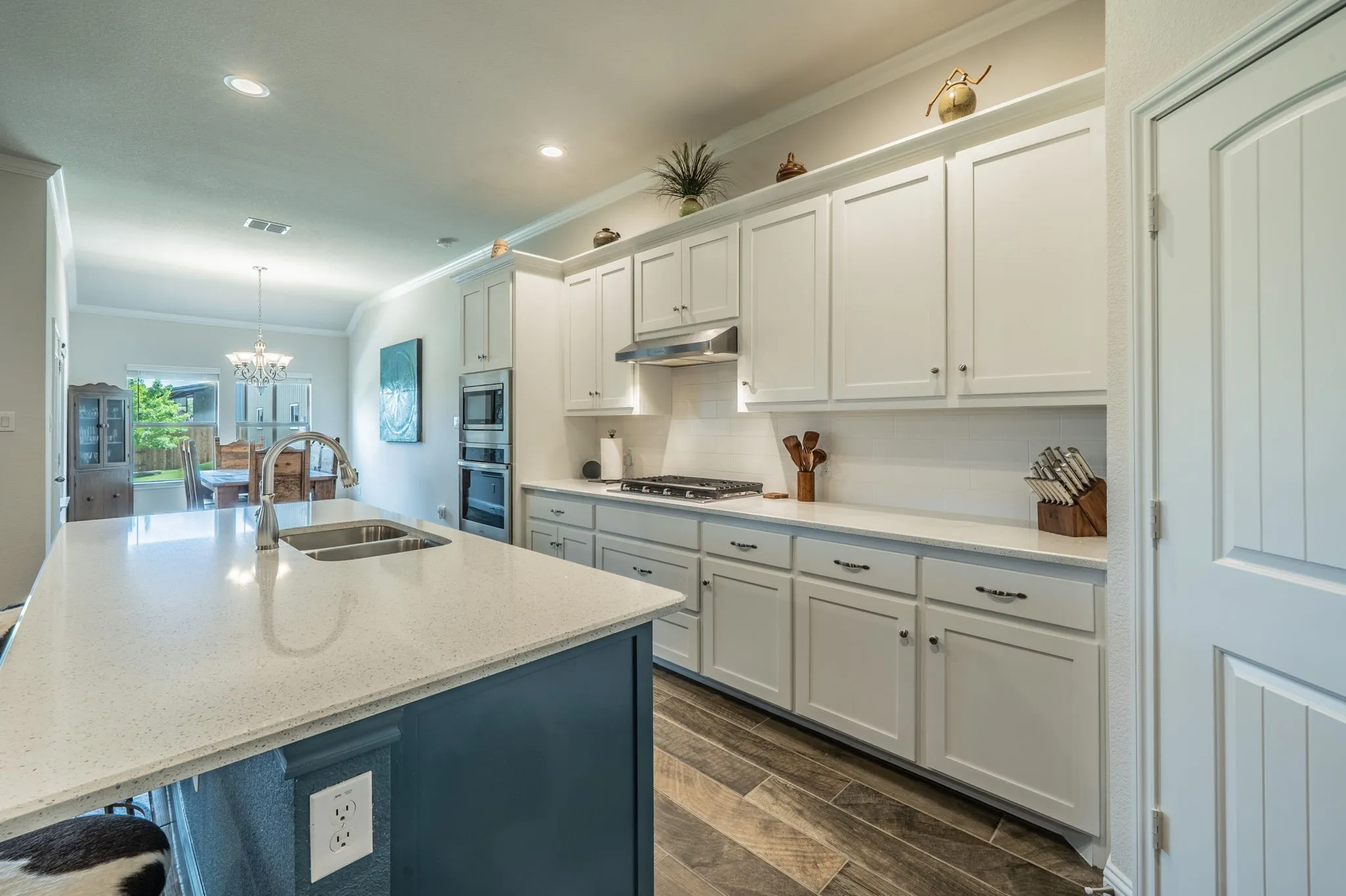 Kitchen featuring white shaker cabinetry, stainless steel appliances, a center island with a built-in sink and dark blue base, light-toned countertops, and wood-finish tile flooring