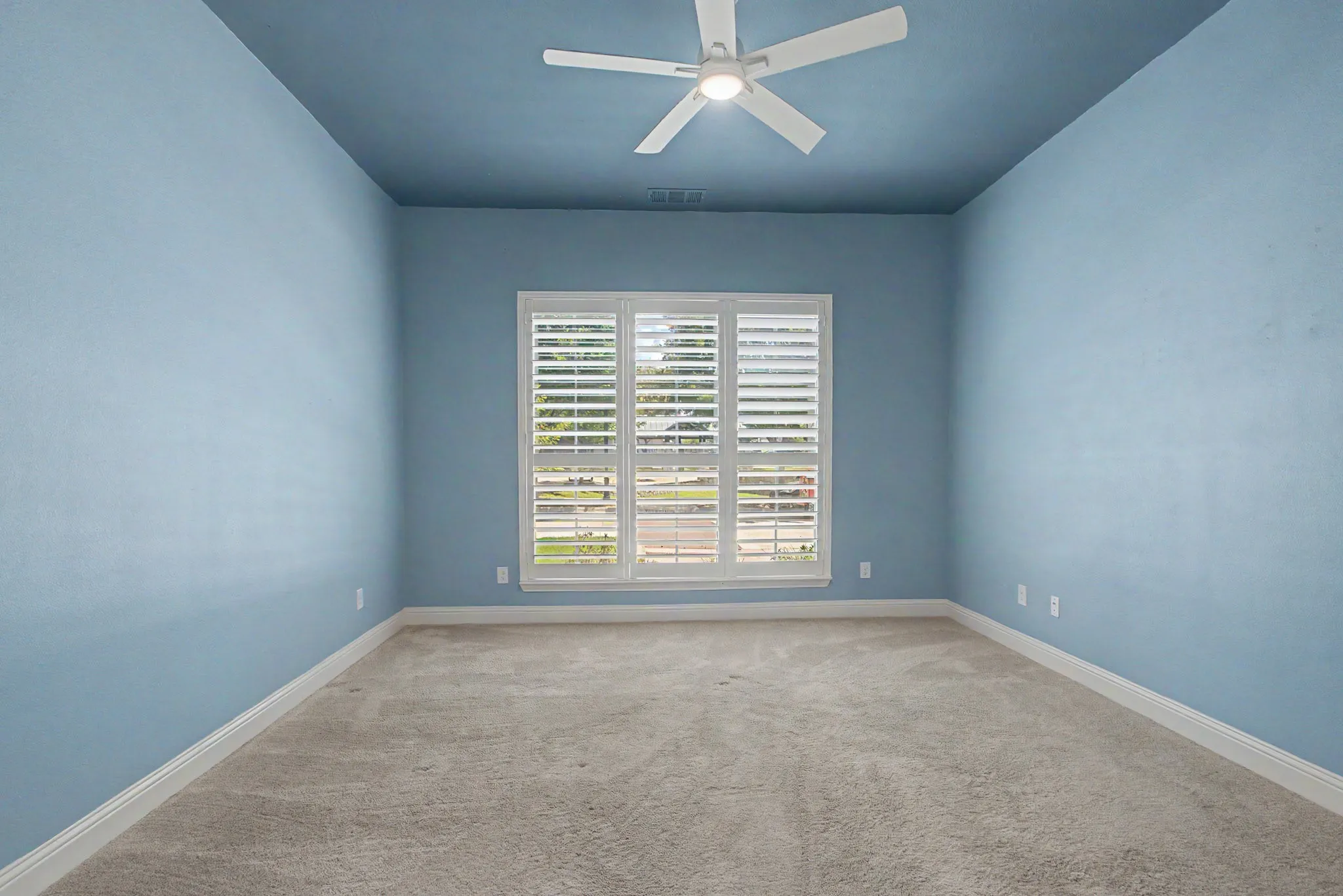 Carpeted room featuring a ceiling fan, three windows with white plantation shutters, and a uniform light blue wall and ceiling finish.Custom Shutters