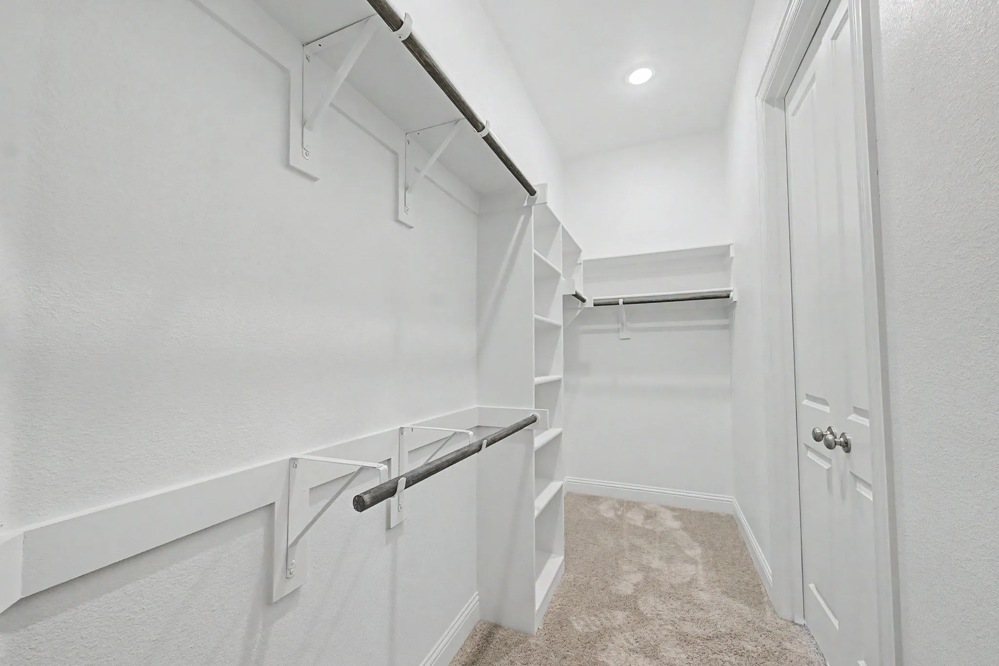 Spacious walk-in closet featuring white built-in shelving, multiple hanging rods, and light-colored carpeting