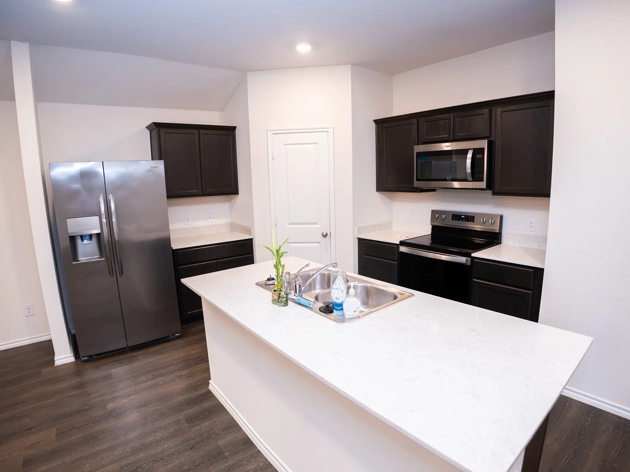 Kitchen featuring dark wood-finish cabinetry, stainless steel appliances, a white solid surface island with an integrated sink, white solid surface countertops, and wood-finish flooring