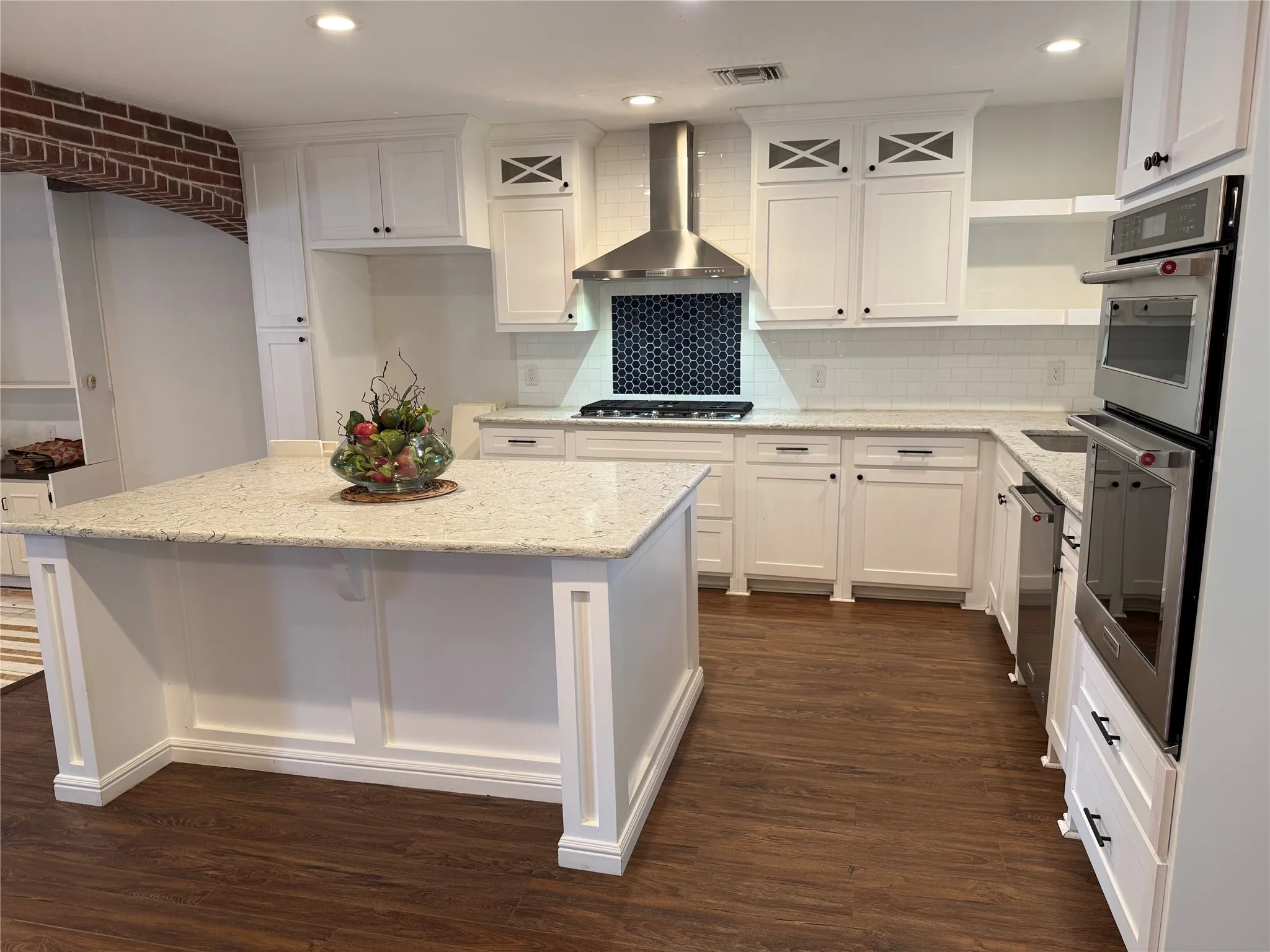 Modern kitchen featuring a central island with a light-toned countertop, white cabinetry with dark hardware, a subway tile backsplash, stainless steel appliances including a built-in double oven, and wood-finish flooring