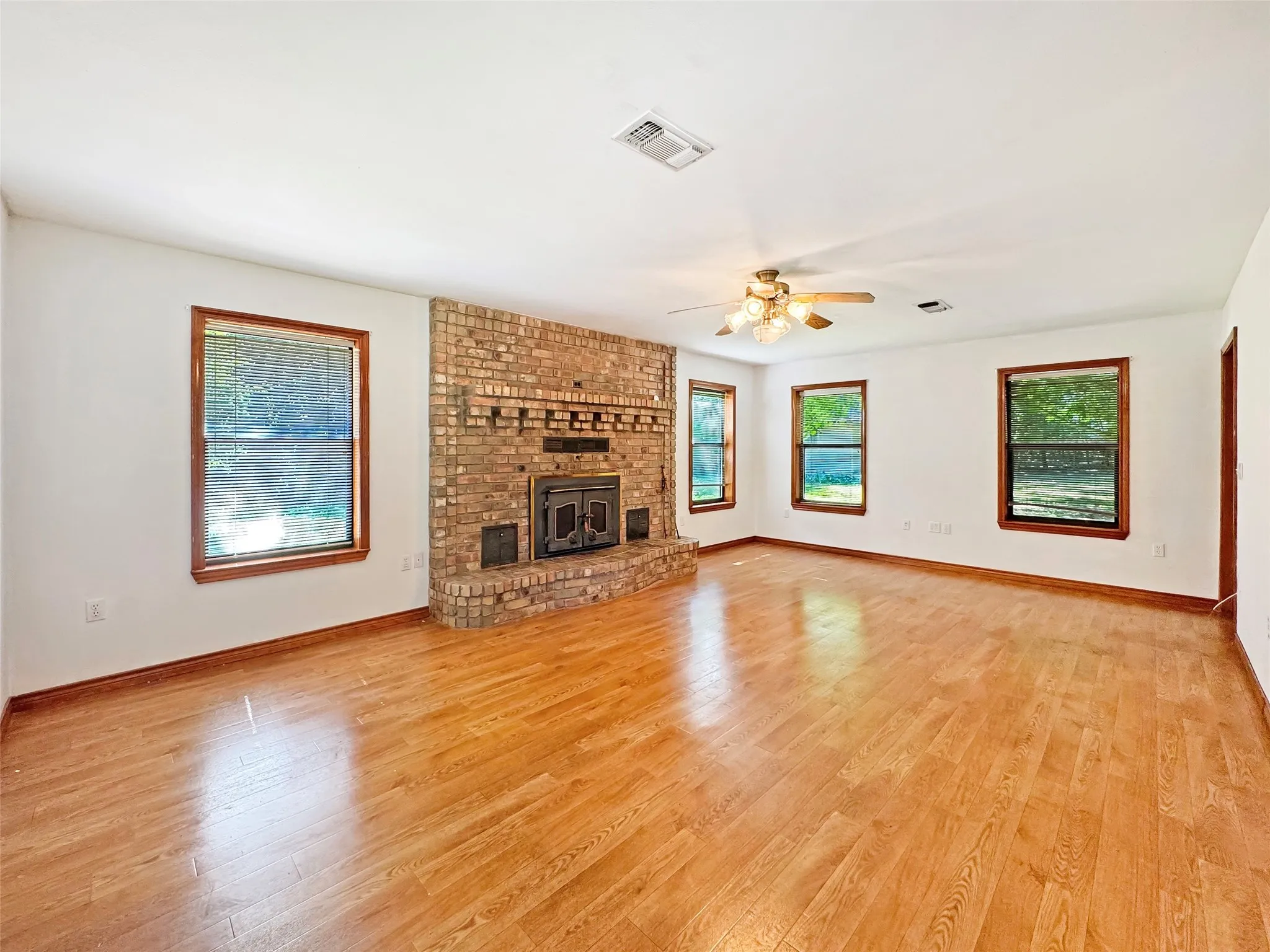 Spacious room featuring a brick fireplace with a raised hearth, wood-finish flooring, multiple windows with wood trim, and a ceiling fan