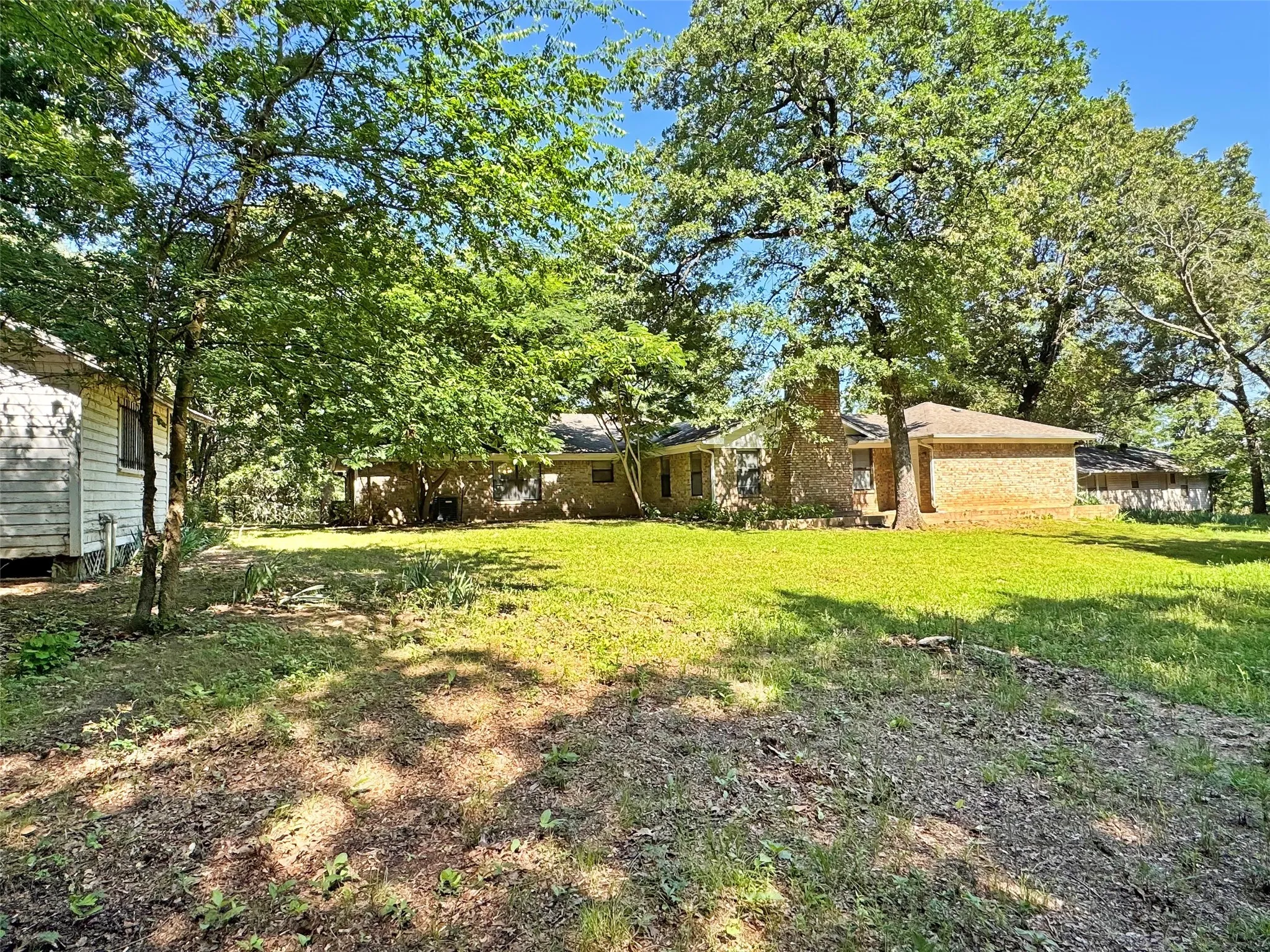 Expansive grassy yard with mature trees and a brick exterior residence featuring a prominent chimney structure
