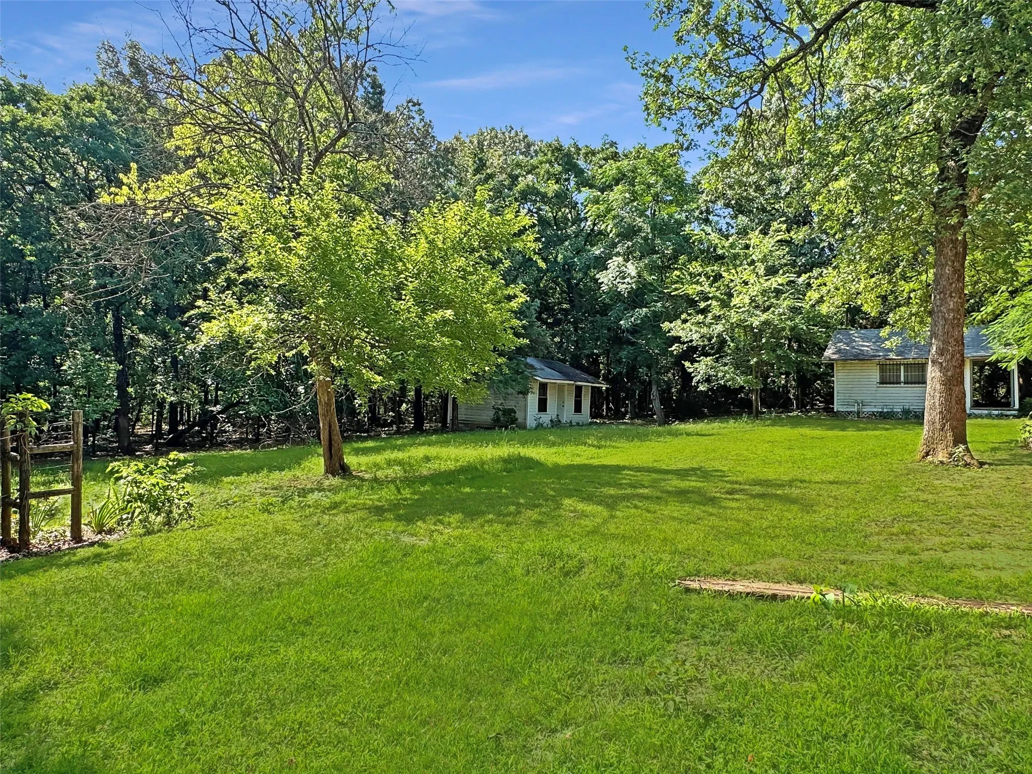 Expansive grassy lawn surrounded by mature trees, featuring two detached outbuildings with light-colored siding and shingled roofs