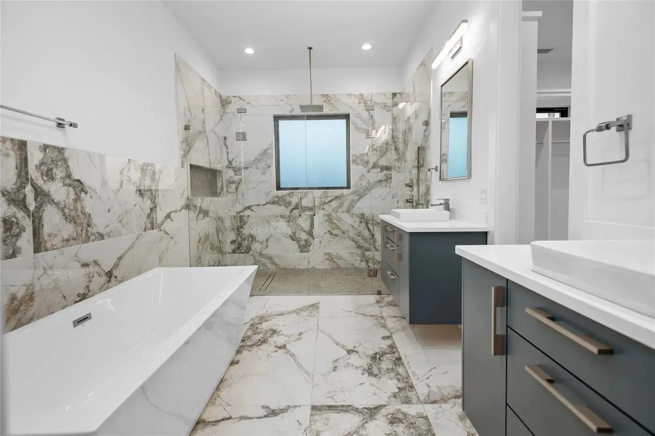 Spacious bathroom featuring large format marble-look tile flooring and walls, a freestanding soaking tub, a frameless glass shower enclosure, and dual vanity sinks with dark cabinetry