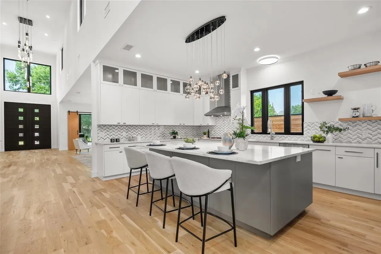 Open-concept kitchen with a large central island, light wood-finish flooring, white cabinetry with glass upper doors, a chevron pattern tile backsplash, and contemporary pendant lighting