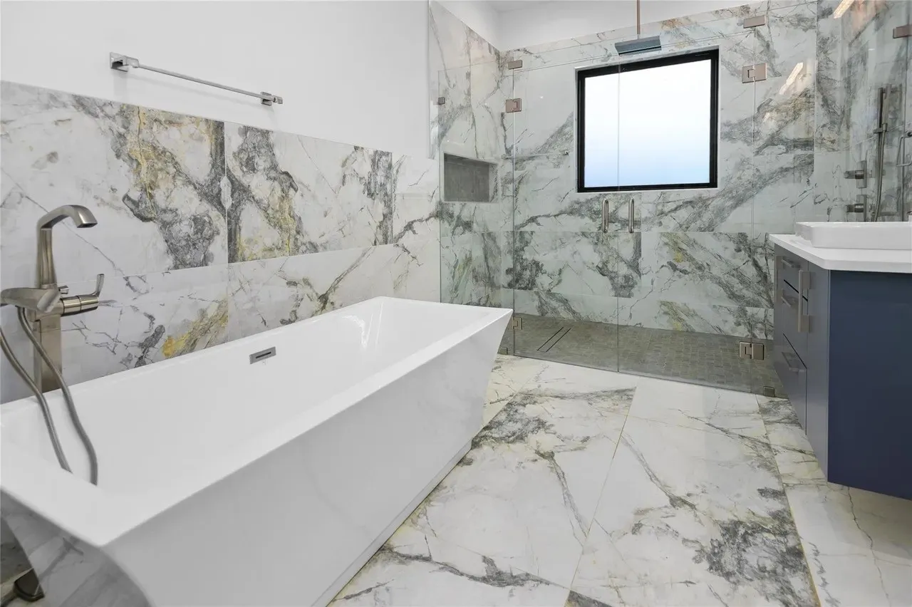 Freestanding soaking tub with a floor-mounted brushed nickel faucet, large format marble-look tile flooring and shower surround, and a glass-enclosed shower with a built-in niche