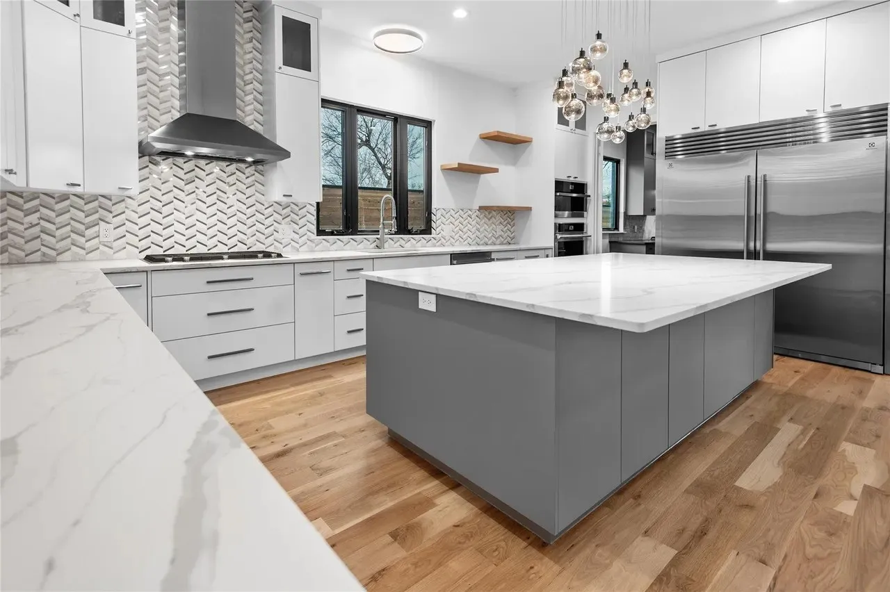 Contemporary kitchen featuring a spacious center island with a white countertop, stainless steel appliances, herringbone pattern backsplash, light wood-finish flooring, and minimalist cabinetry