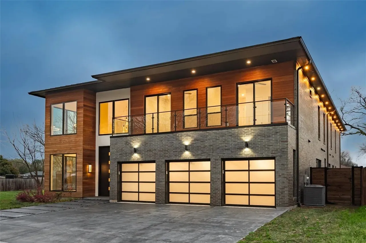 Modern exterior featuring wood paneling, dark brick accents, a three-car garage with frosted glass doors, a second-story balcony with glass railing, and a concrete drive
