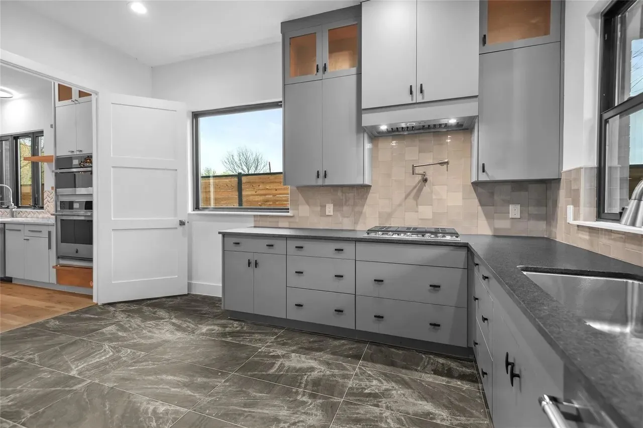 Modern kitchen featuring stone-finish floor tiling, grey cabinetry with dark hardware, dark countertops, and a tiled backsplash
