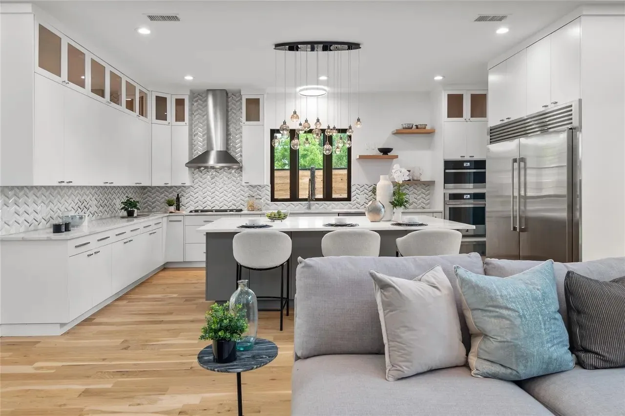 Open-concept kitchen featuring an oversized island with seating, a chevron-patterned tile backsplash, stainless steel appliances, abundant white cabinetry with glass upper accents, and wood-finish flooring