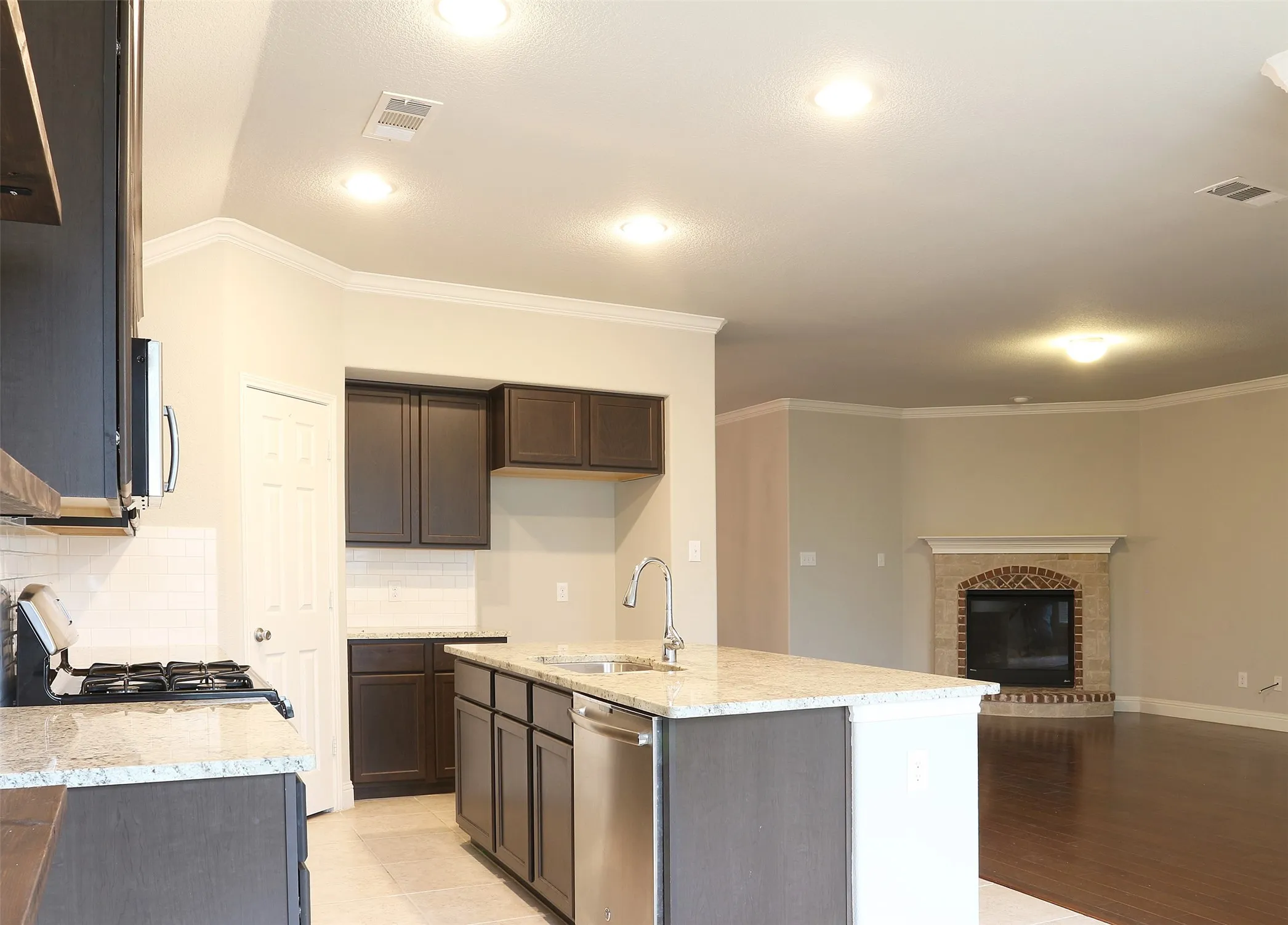 Open-concept kitchen and living area featuring a central island with a stainless steel sink and appliance, dark wood-finish cabinetry, light-toned countertops, and a brick fireplace