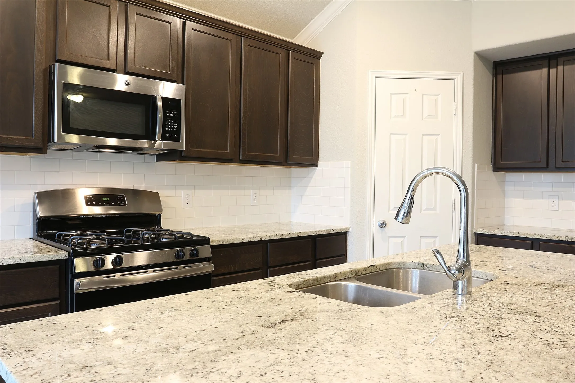 Kitchen featuring dark wood-finish cabinetry, stainless steel appliances including a gas range and built-in microwave, light-toned speckled countertops, a double basin sink with a chrome faucet, and white subway tile backsplash