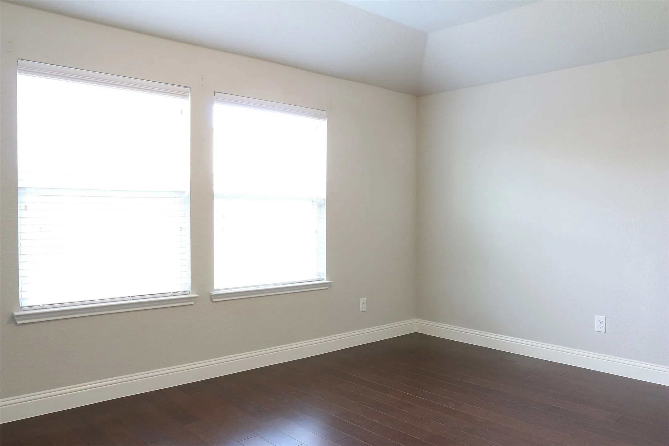 Room featuring two windows with horizontal blinds, rich wood-finish flooring, white baseboards, and a tray ceiling