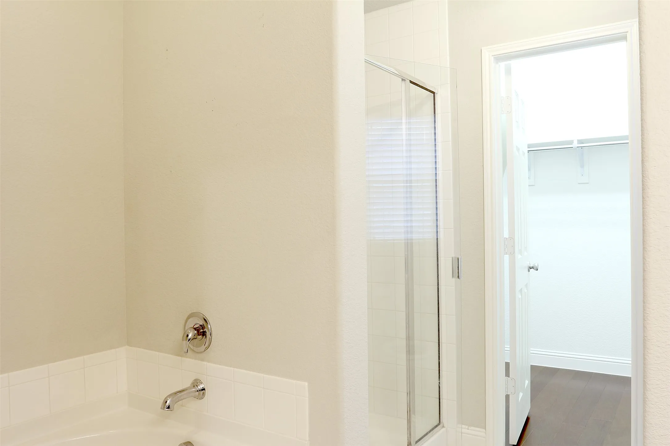 Bathtub with polished chrome fixtures, glass-enclosed shower, white tile accents, and a separate closet with wood-finish flooring and built-in shelving