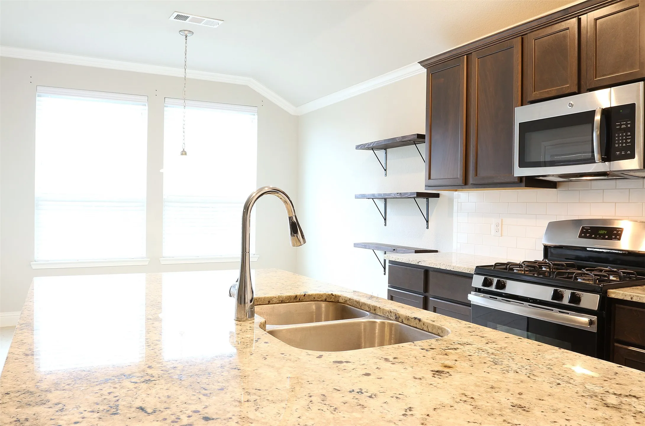 Kitchen island featuring a double basin stainless steel sink with a high-arch faucet, granite countertop, dark wood-finish cabinetry, stainless steel microwave, and gas range