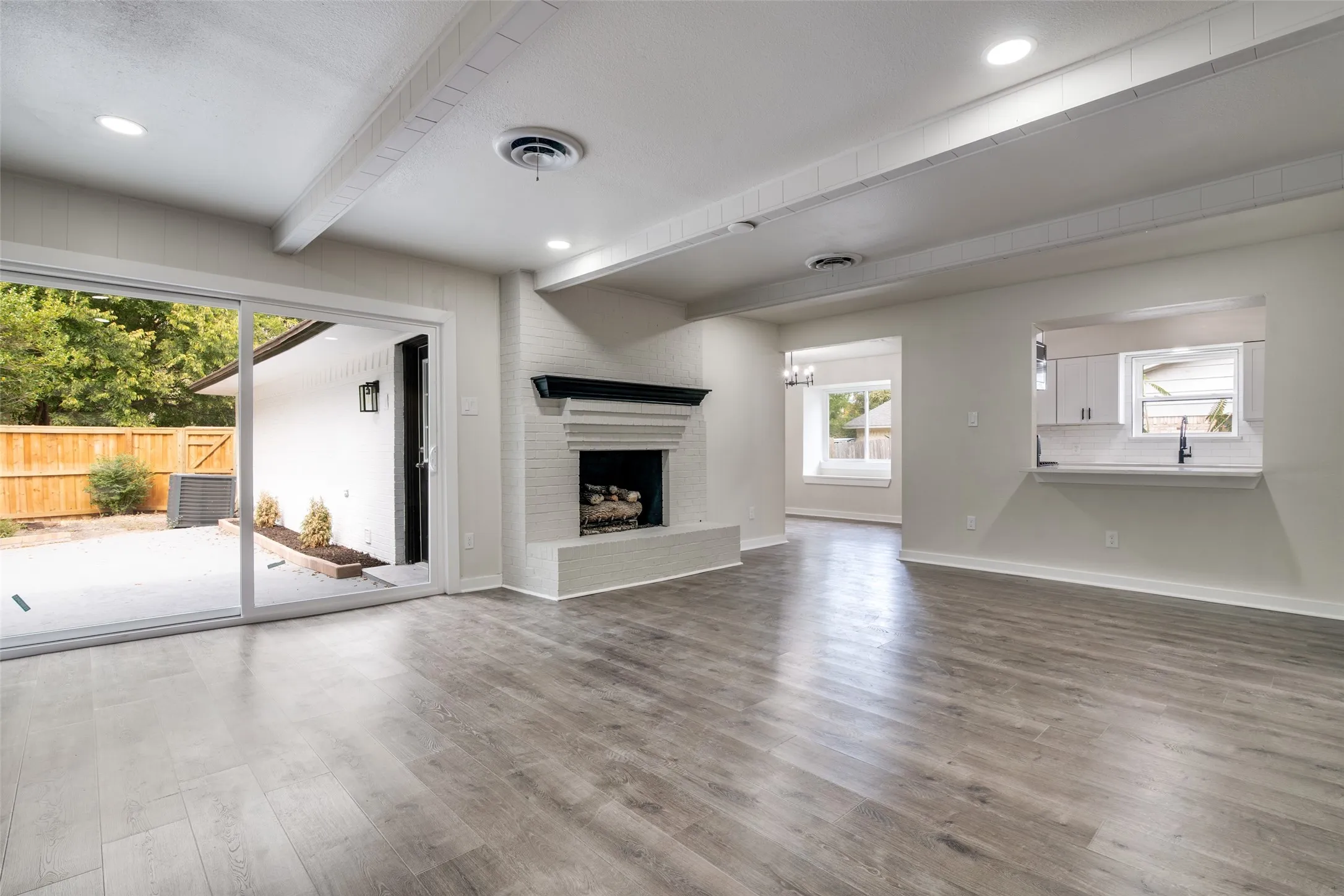 Spacious living area featuring wood-finish flooring, a white brick fireplace with a black mantel, and exposed ceiling beams