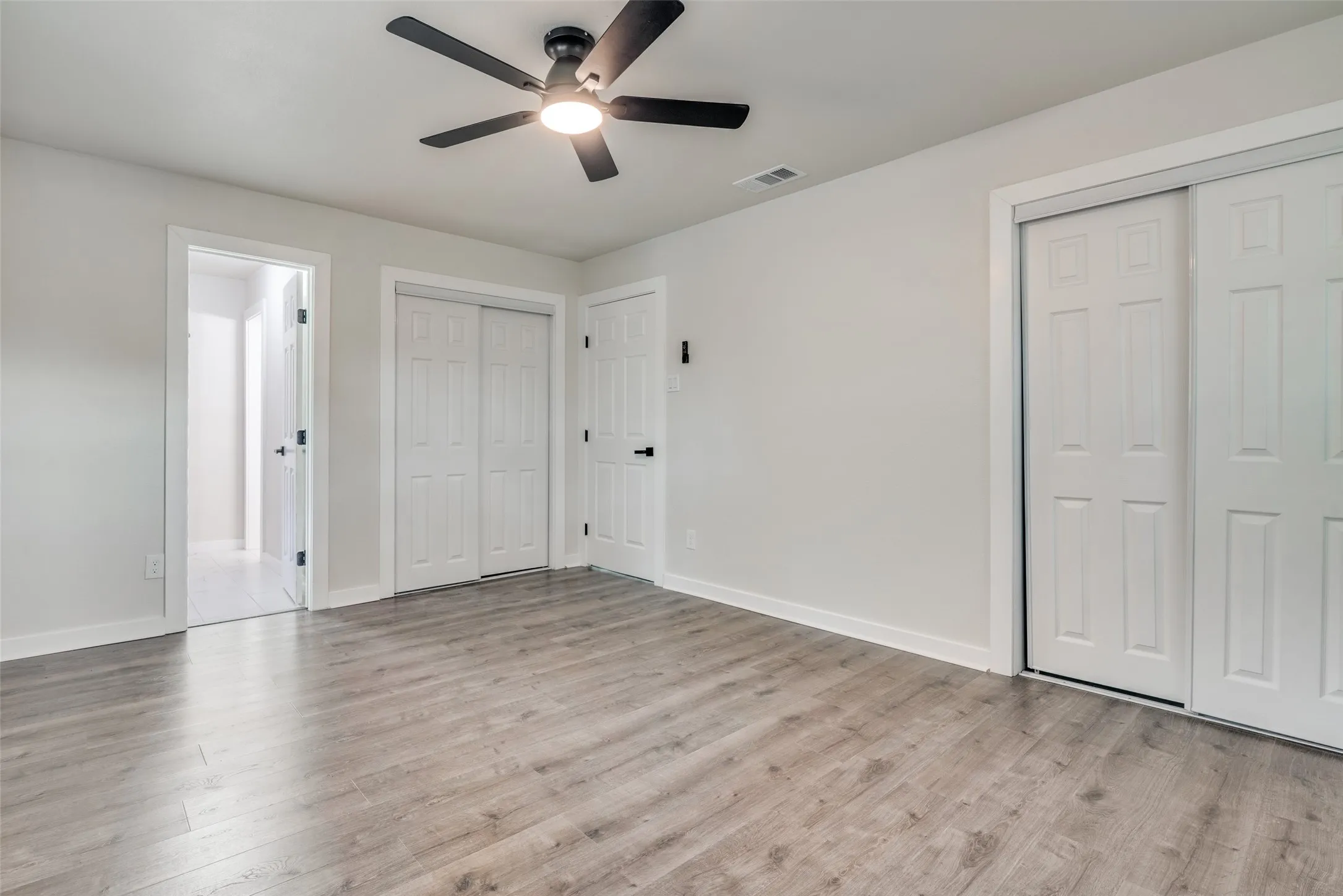 Spacious room featuring wood-finish flooring, light gray wall tones, a ceiling fan with integrated lighting, and multiple doors with contrasting black hardware