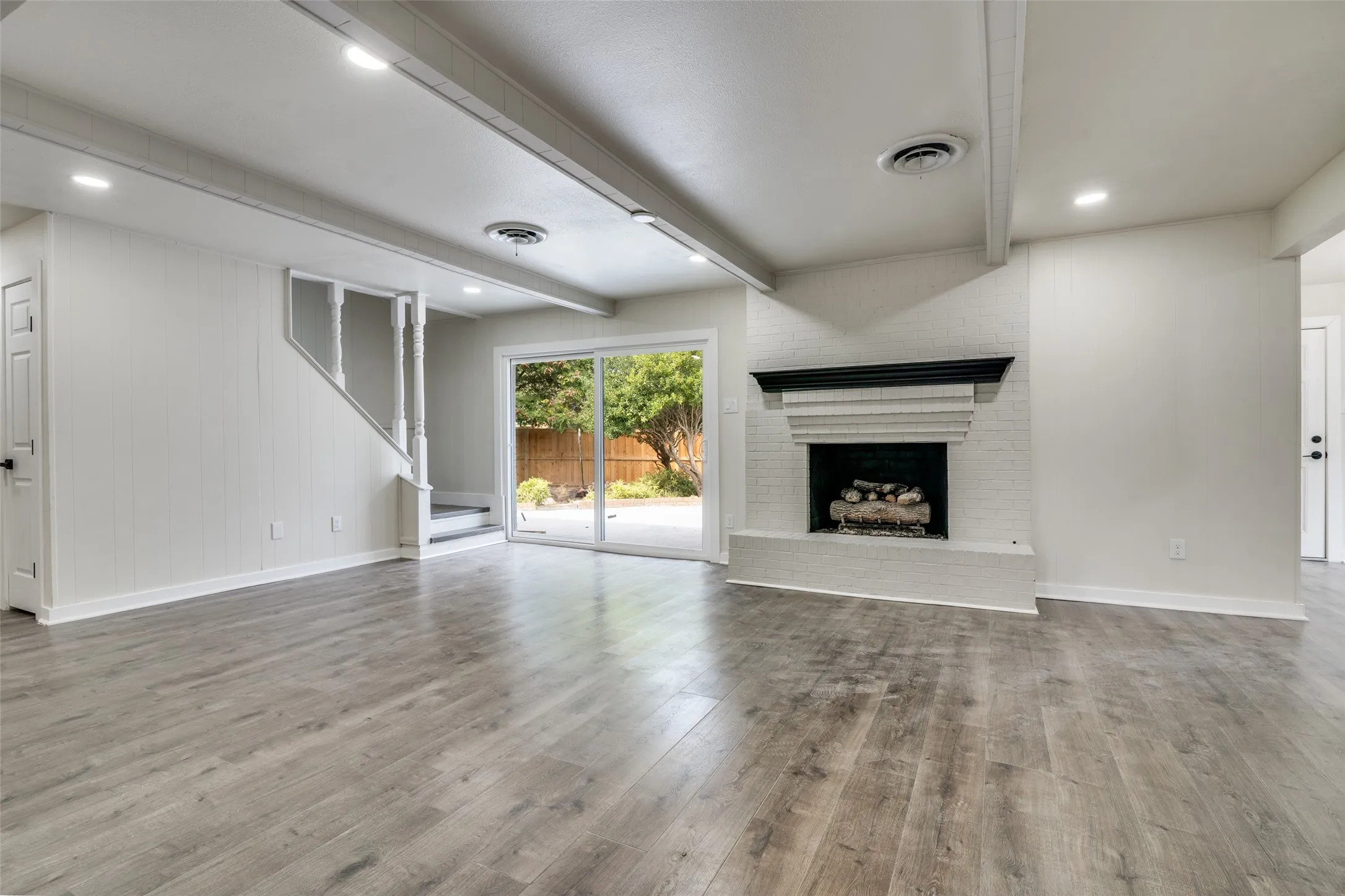 Spacious room featuring wood-finish flooring, a white-painted brick fireplace with a dark mantel, a staircase with white balusters, recessed lighting, and sliding glass doors leading to exterior space