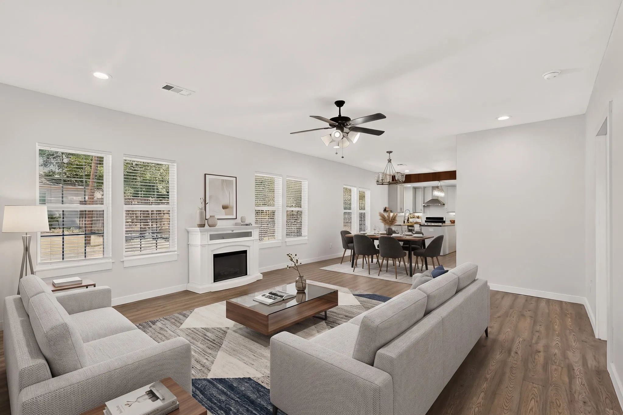 Open-concept living space with wood-finish flooring, a white fireplace surround, multiple windows, recessed lighting, and a ceiling fan