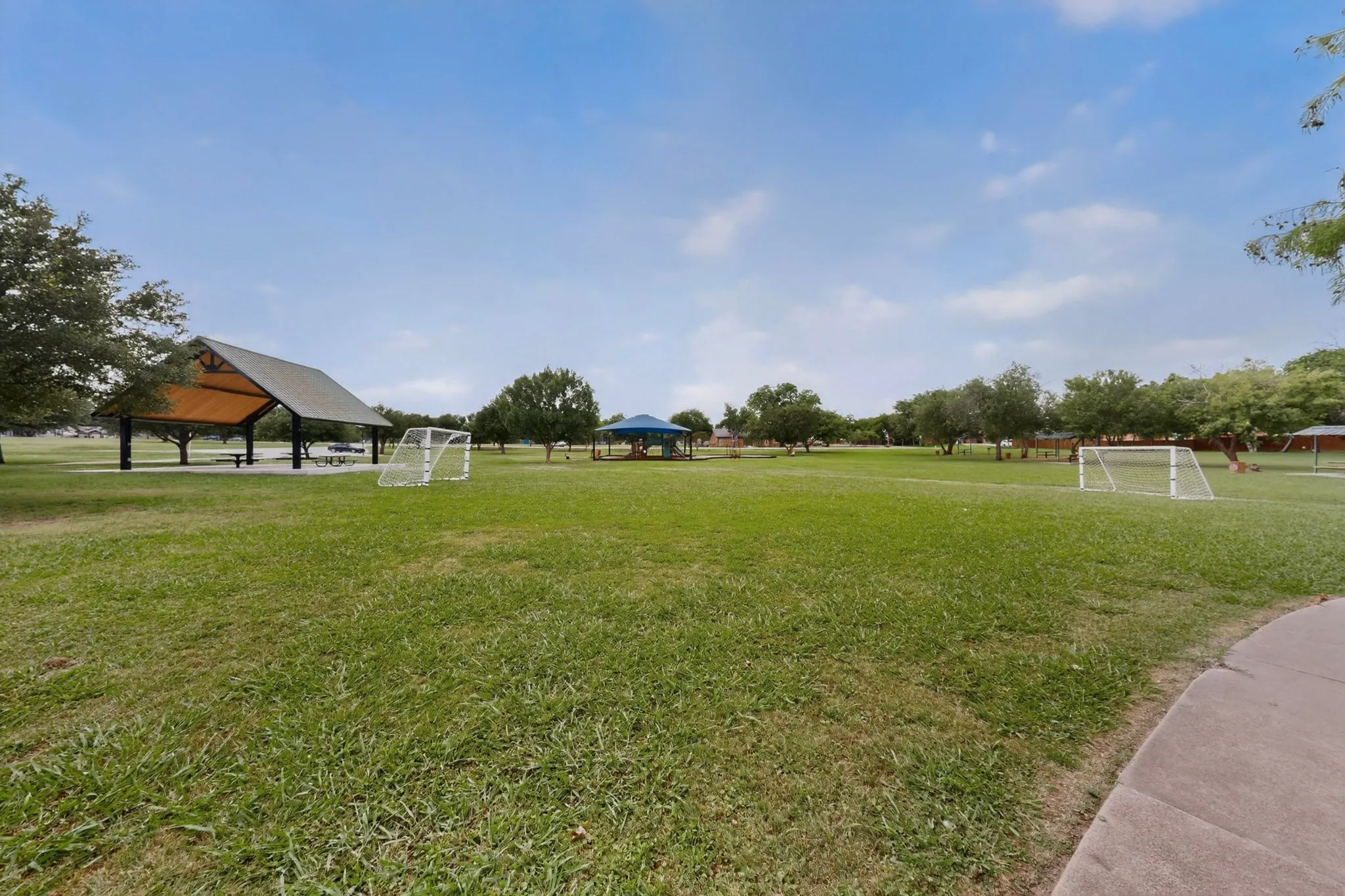Expansive green space featuring two soccer goals, a large covered picnic area, mature trees, and a paved walking path