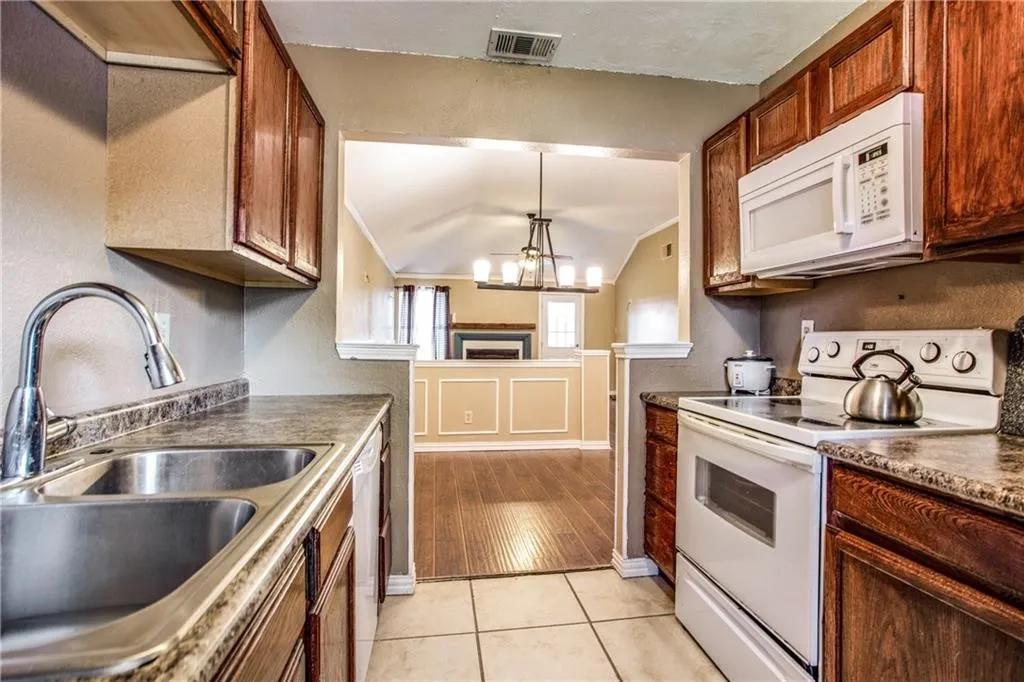Kitchen featuring a double stainless steel sink with a gooseneck faucet, white electric range, white built-in microwave, wood-finish cabinetry, and tiled flooring