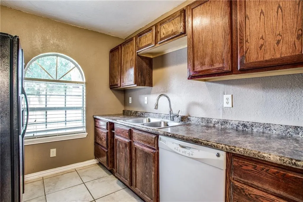 Kitchen featuring rich wood cabinetry, patterned countertops, a double basin sink with a gooseneck faucet, an arched window, and tile flooring