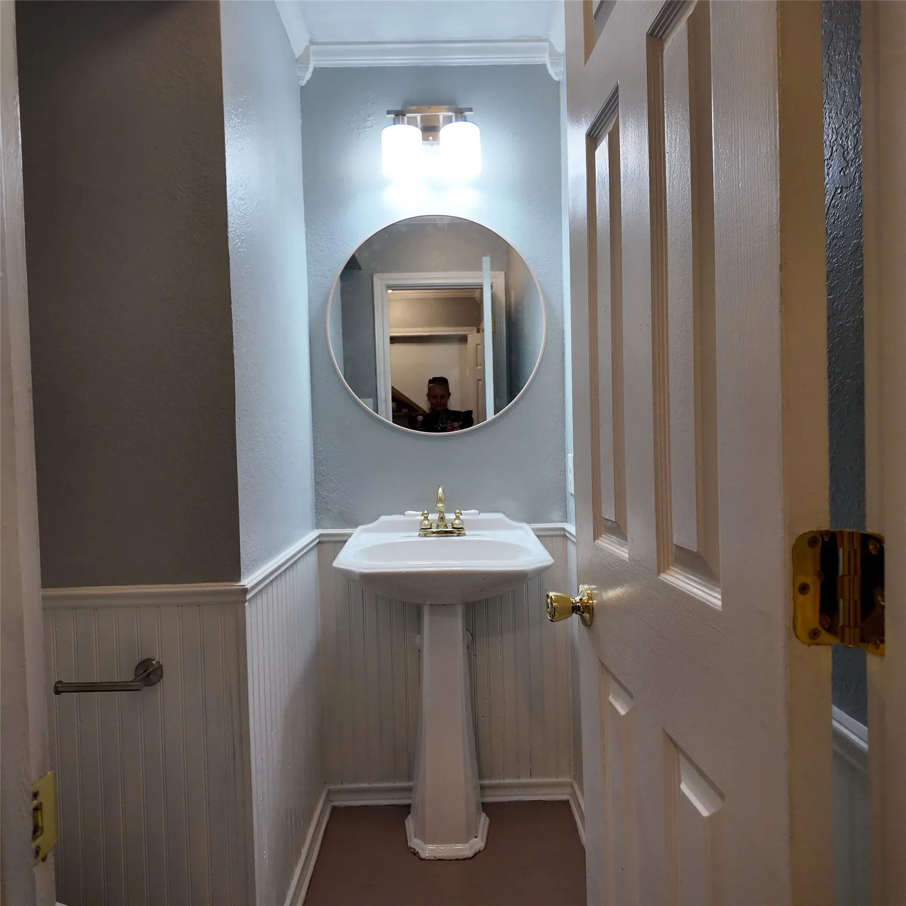 Pedestal sink with brass-finish faucet, circular wall mirror, and wainscoting