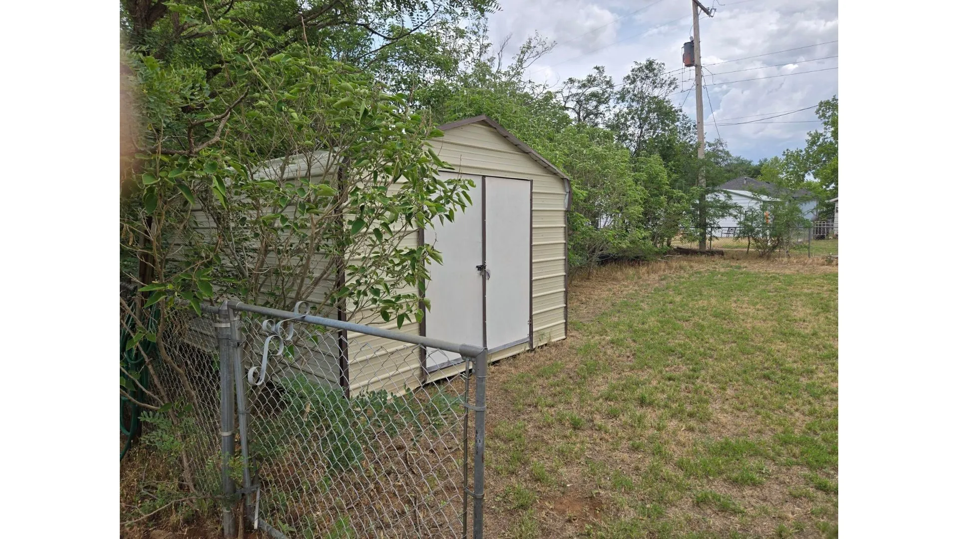 Metal utility shed with double panel doors, situated in a grass area with mature greenery and a chain link fence