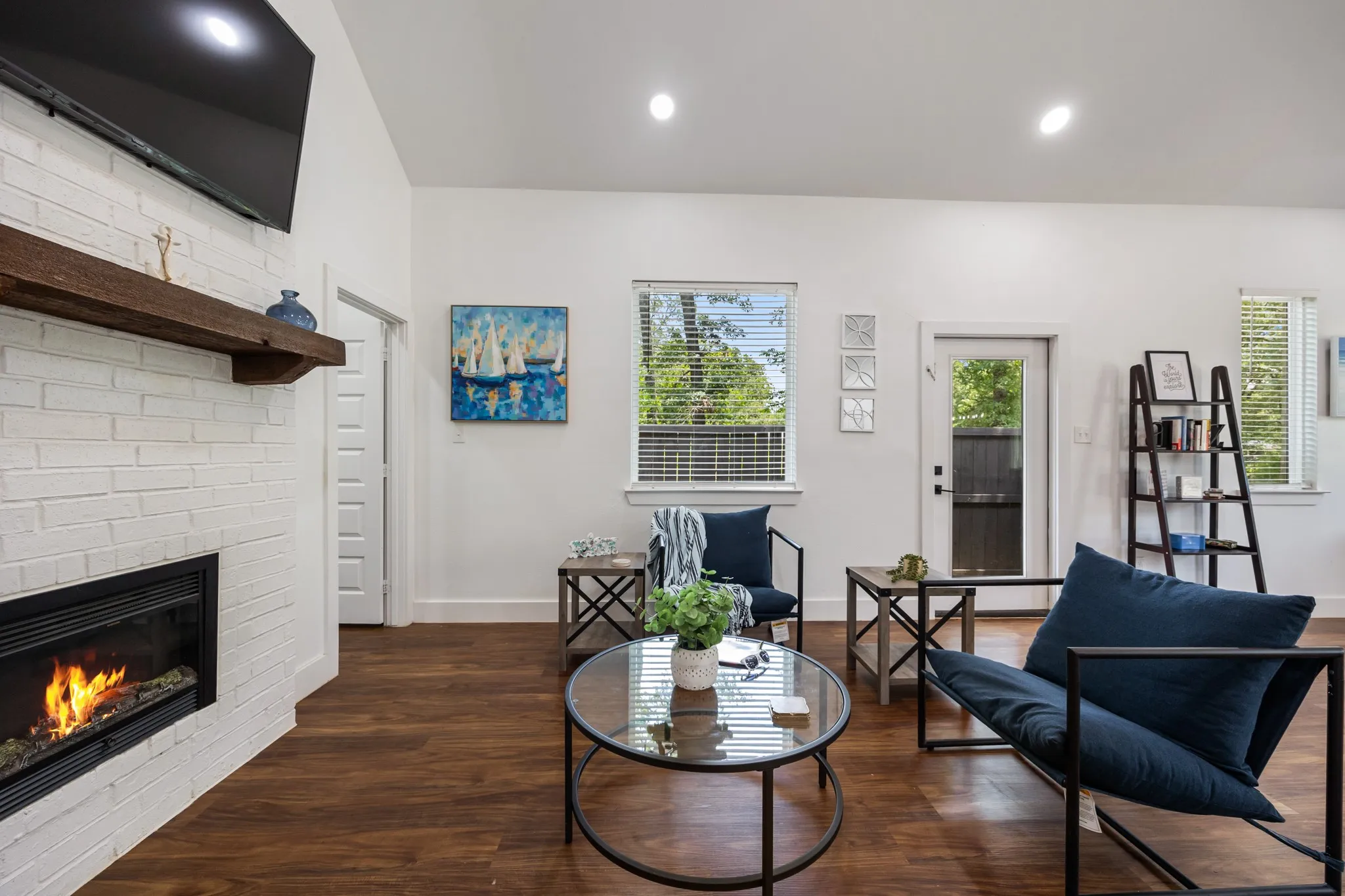 Living area featuring wood-finish flooring, a white brick fireplace with a wood mantle, recessed lighting, and multiple windows with horizontal blinds