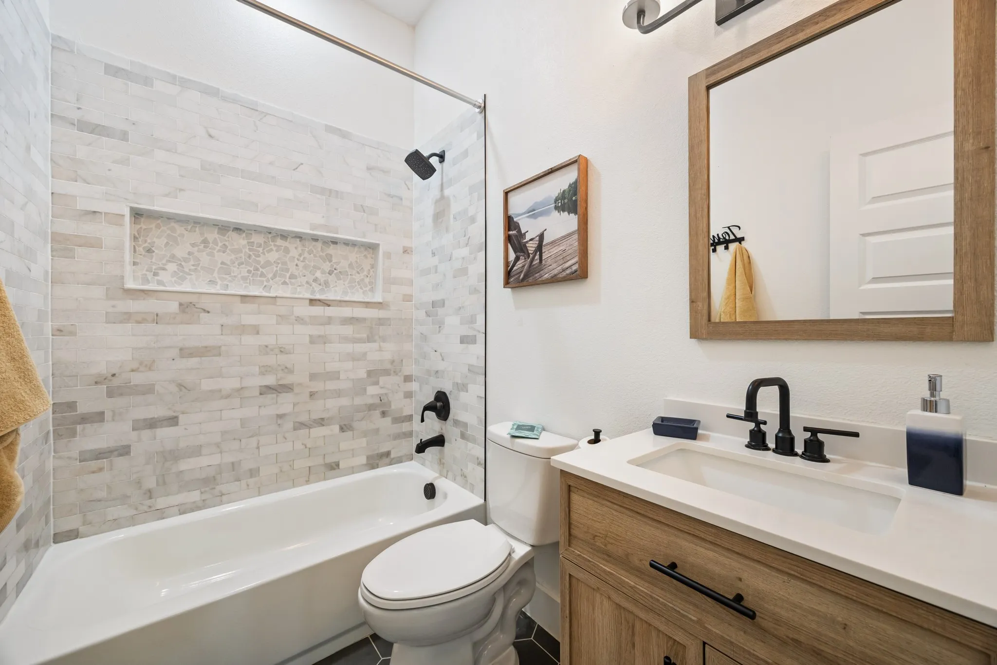 Bathroom featuring a tiled shower/tub combination with a recessed niche, a wood-finish vanity with a white countertop and integrated sink, a rectangular mirror with a wood-finish frame, and matte black plumbing fixtures