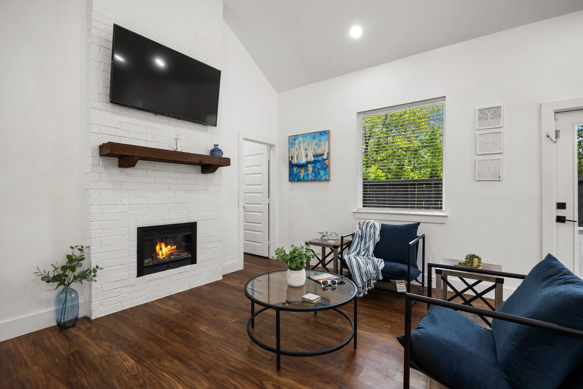 Vaulted ceiling living area featuring a white painted brick fireplace with a dark wood mantel, wood-finish flooring, and a large window