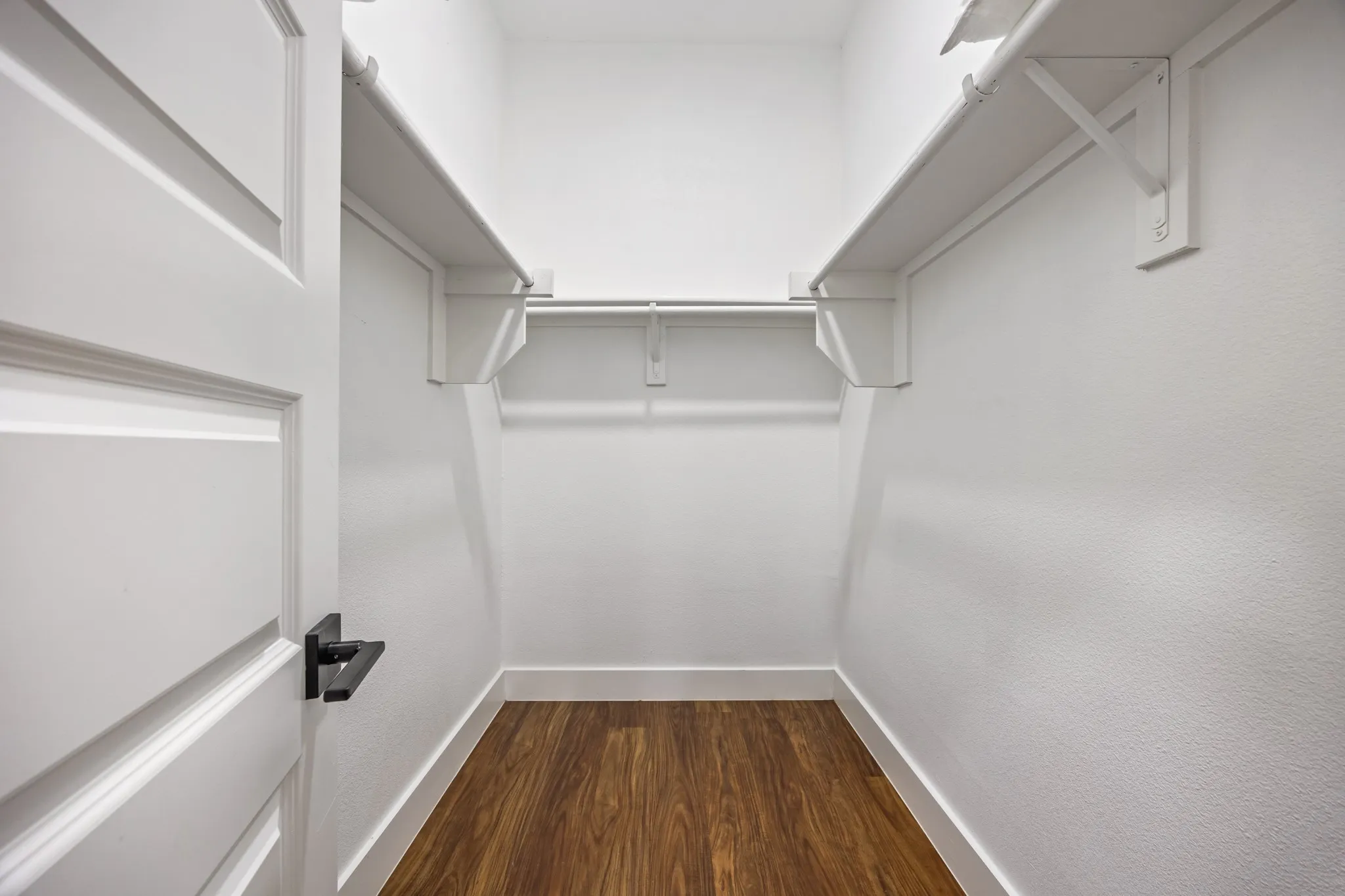 Walk-in closet featuring wood-finish flooring, white walls, white shelving, a clothing rod, and a white door with a black lever handle