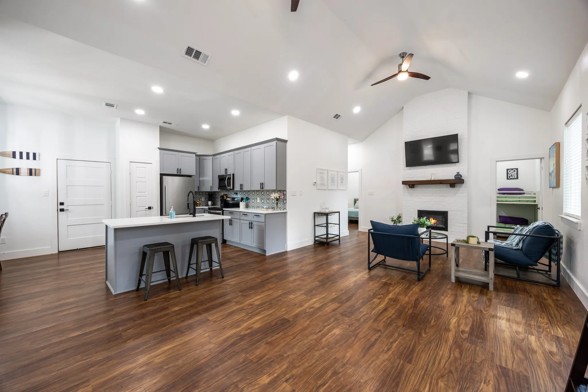 Open-concept interior featuring wood-finish flooring, a kitchen island with seating, stainless steel appliances, a white brick fireplace, and vaulted ceilings