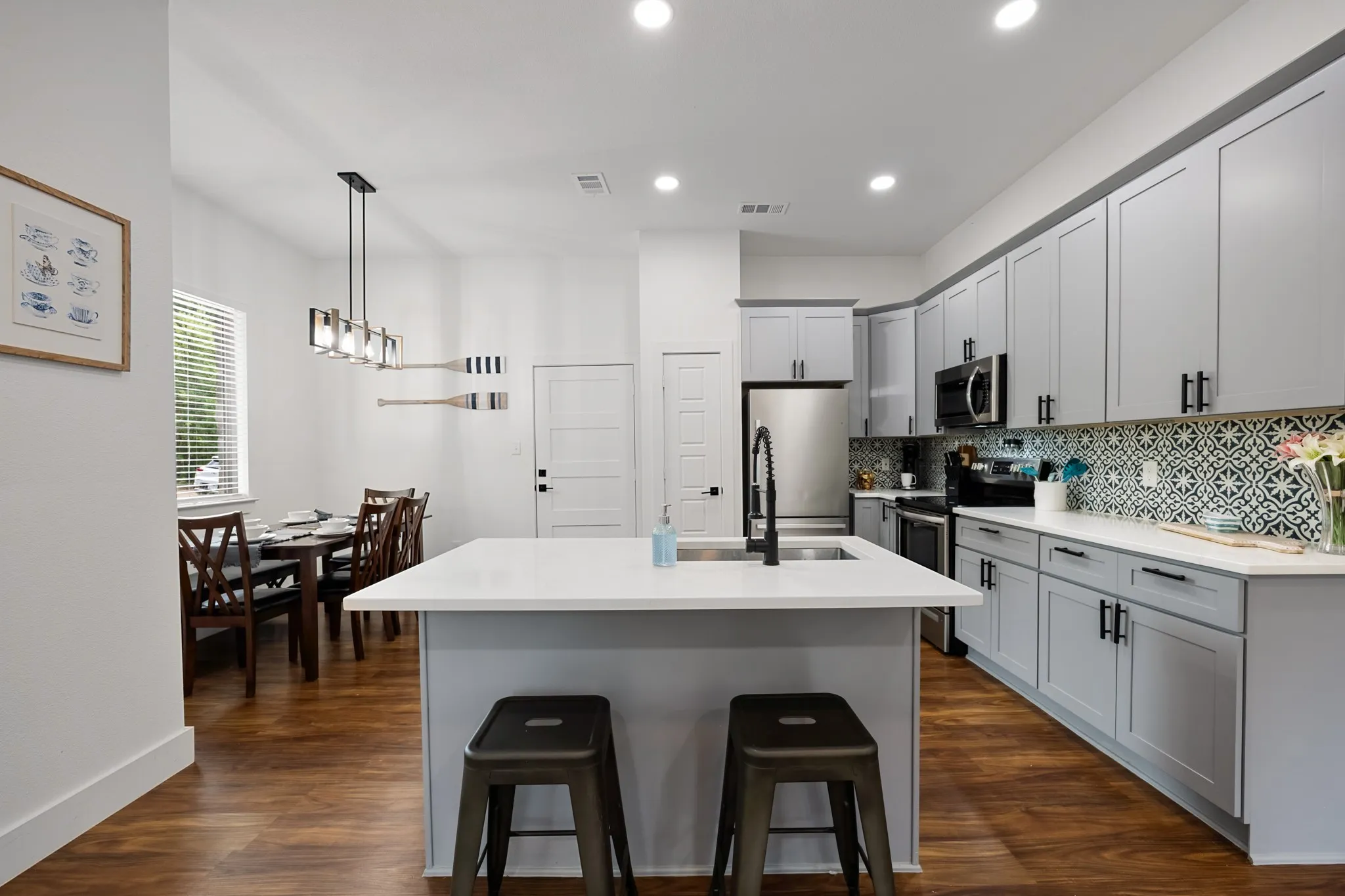 Open-concept kitchen featuring an island with a built-in sink, stainless steel appliances, light gray cabinetry with matte black hardware, white countertops, and a patterned tile backsplash