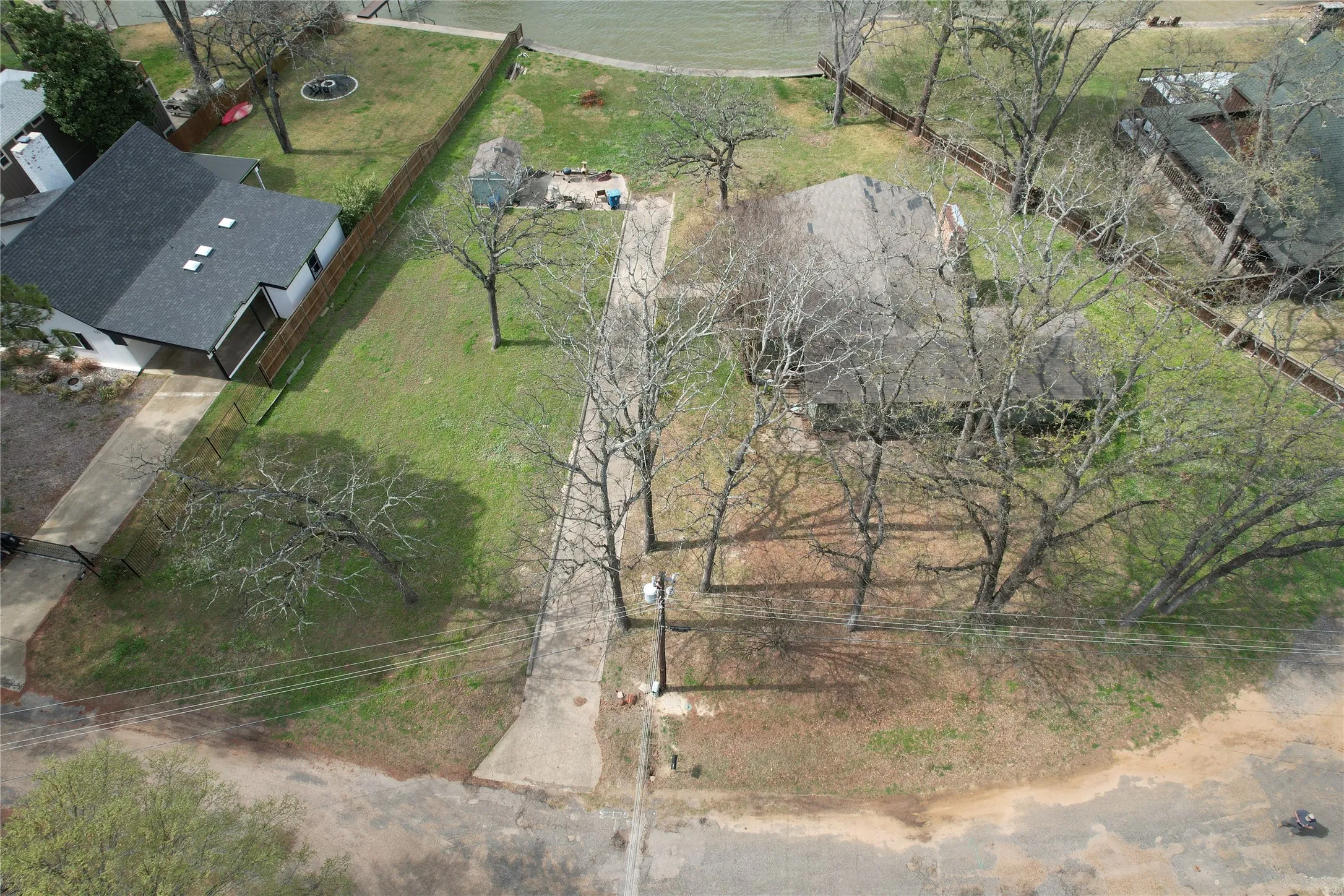 Aerial view from Street to Lake of 135 Island Park Drive showing the fenced property side property lines, Island Park Drive in the foreground and Cedar Creek Lake front in the background, and the long paved driveway providing access to a house and land featuring mature trees, a detached storage shed, and a open water view to the west