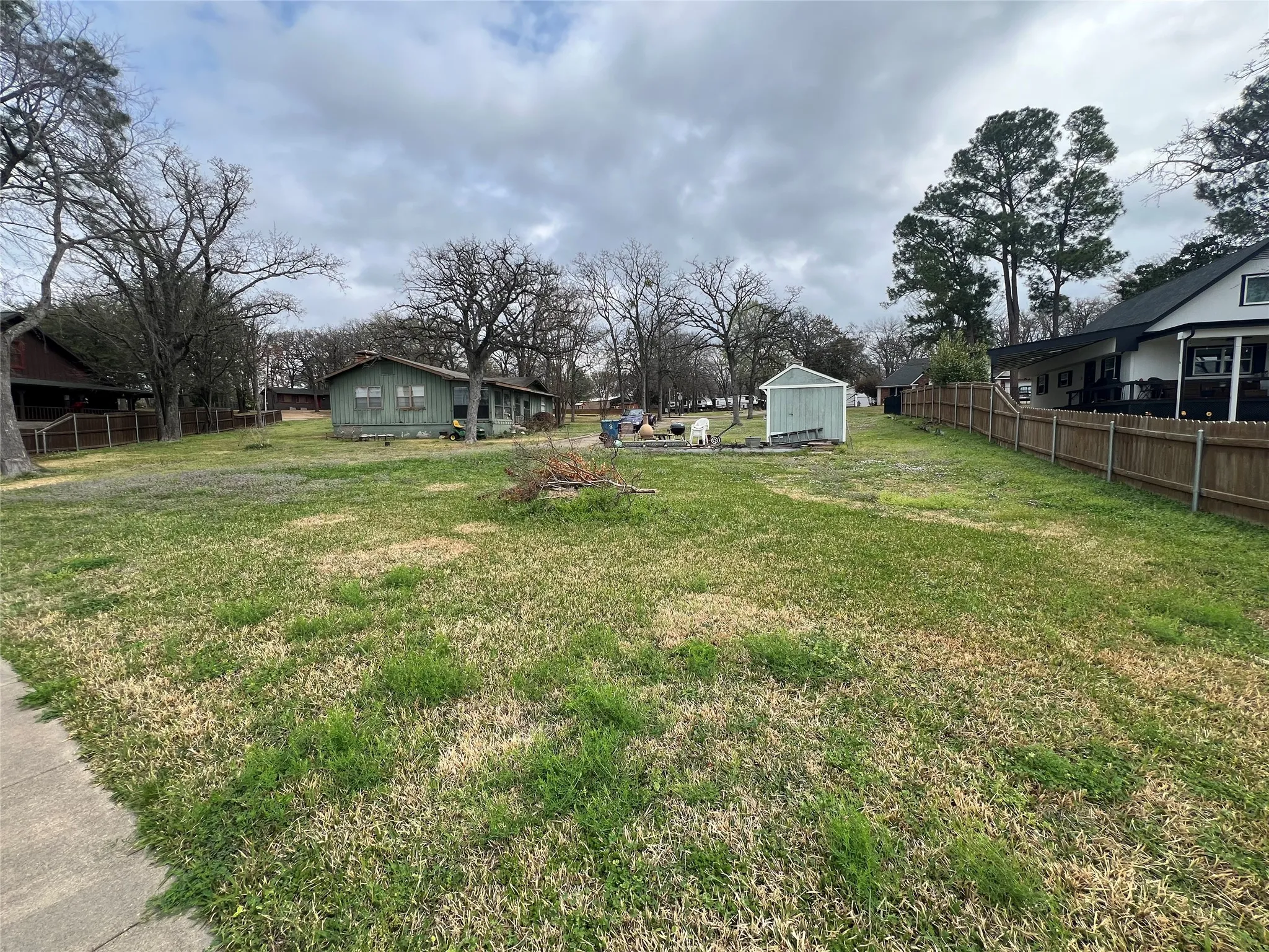 Expansive grassy lot featuring a light green storage shed, established trees, and a wood privacy fence