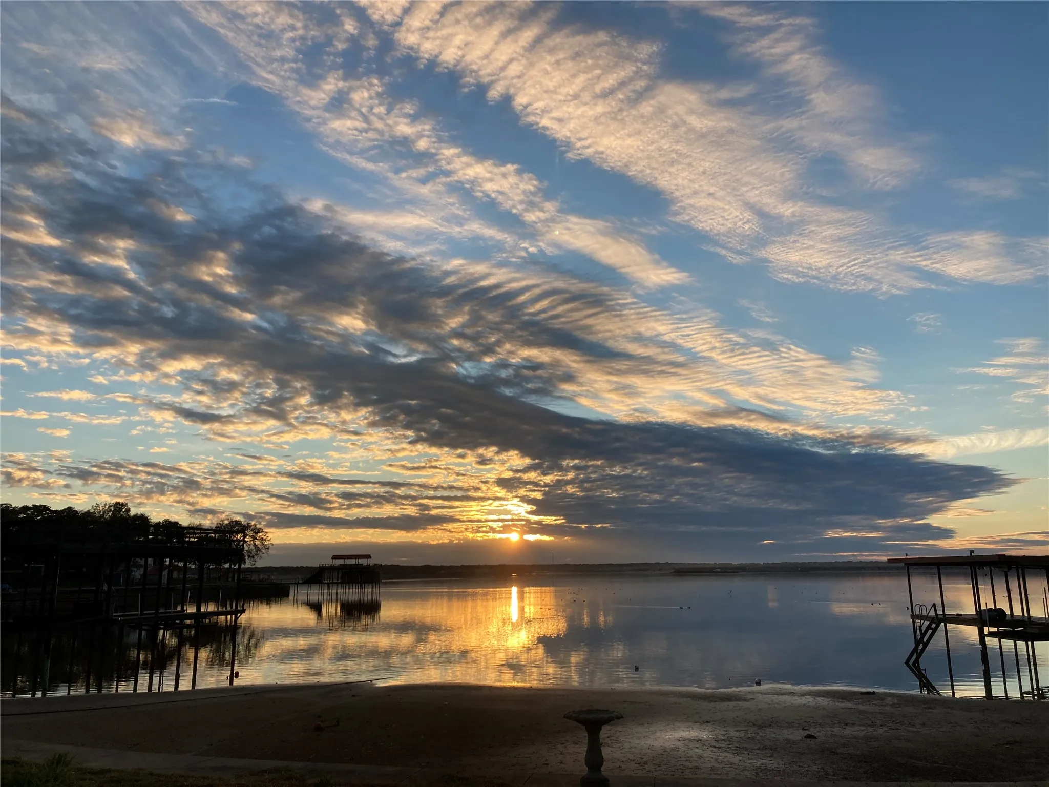 Waterfront setting featuring a sandy shoreline with a bird bath, calm water with reflections, and distant docks with covered boat slips