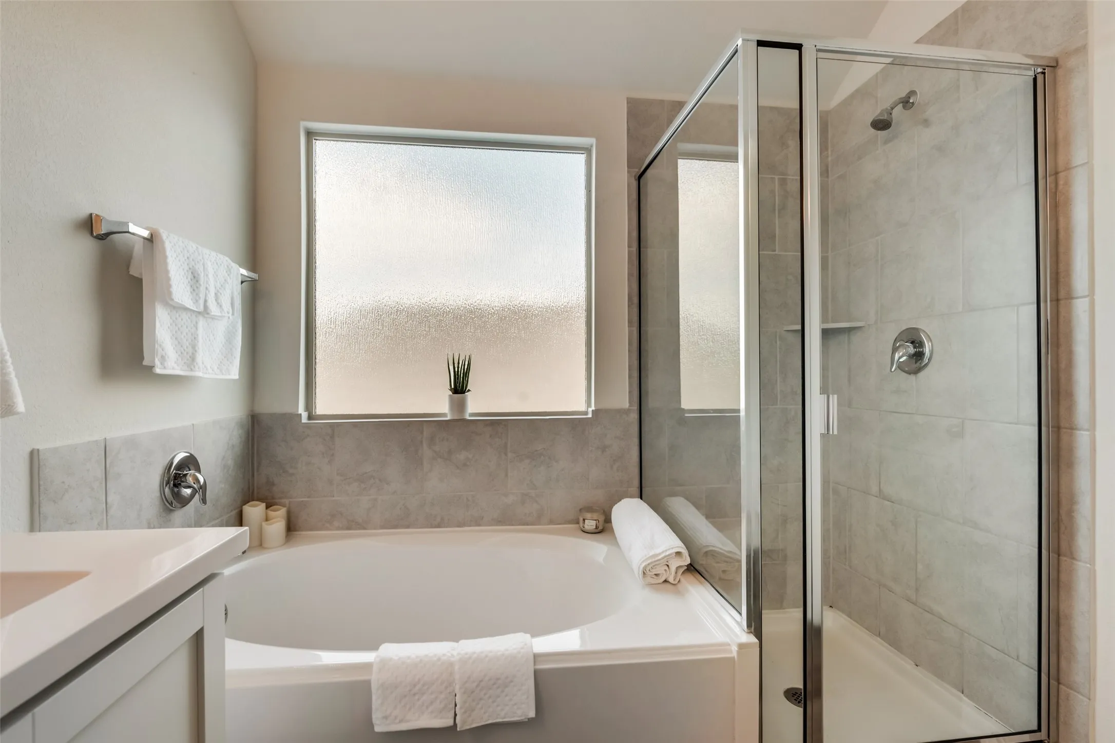 Bathroom featuring a built-in soaking tub with a tiled surround, a separate glass-enclosed shower with tile walls, a frosted privacy window, and a vanity with a light-toned countertop